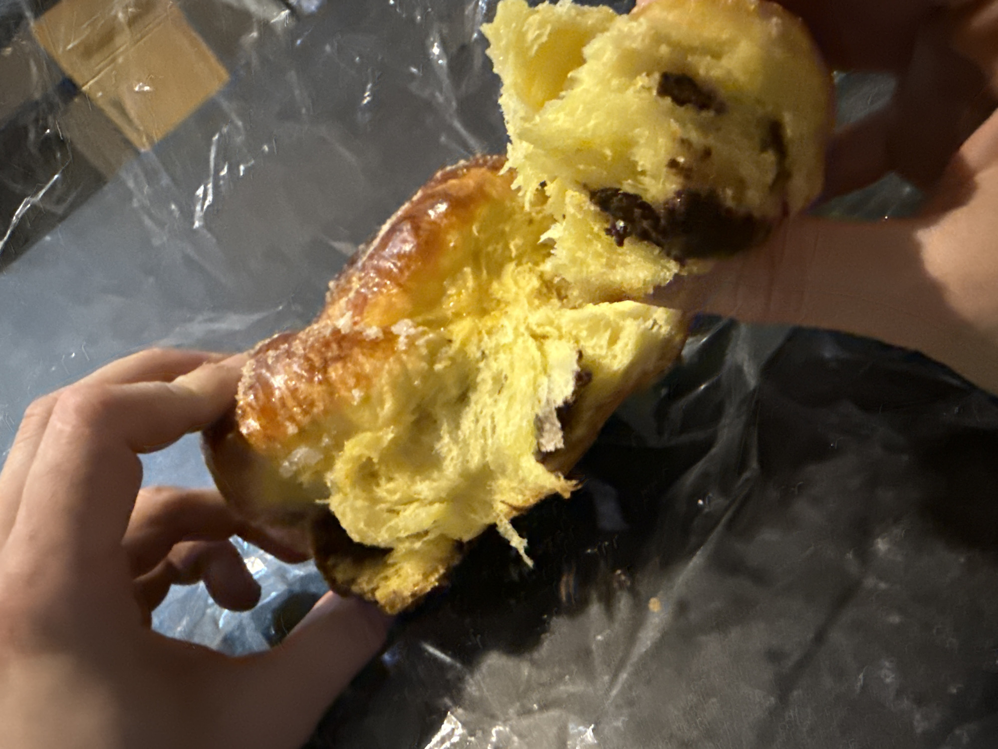 A person tears a pastry in half with their hands, revealing a yellow interior with a dark filling. The pastry has a glazed, golden-brown crust. The pastry and hands are resting on crinkled plastic wrap. A portion of a box is visible in the background behind the plastic wrap.