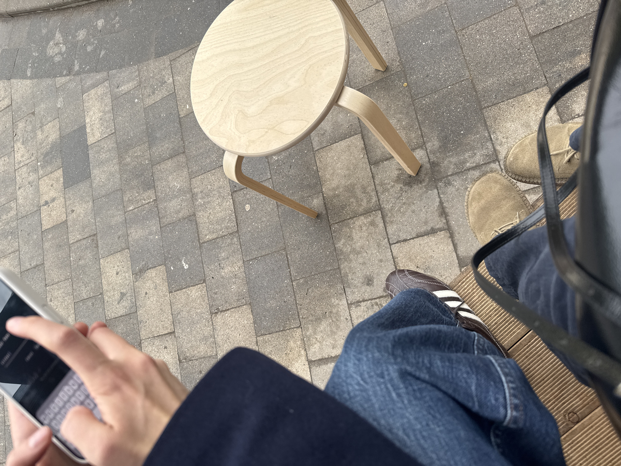 The image depicts a person sitting outdoors on a wooden bench, using a smartphone. Another person stands or sits nearby. There is a light-colored, wooden, round stool next to the bench. The ground is paved with grey blocks. The person using the phone wears a dark coat and jeans, and one shoe is visible, a brown sneaker with white stripes. The other person wears tan shoes and jeans. The location is most likely a public outdoor seating area in an urban environment, potentially Moscow, Russia.