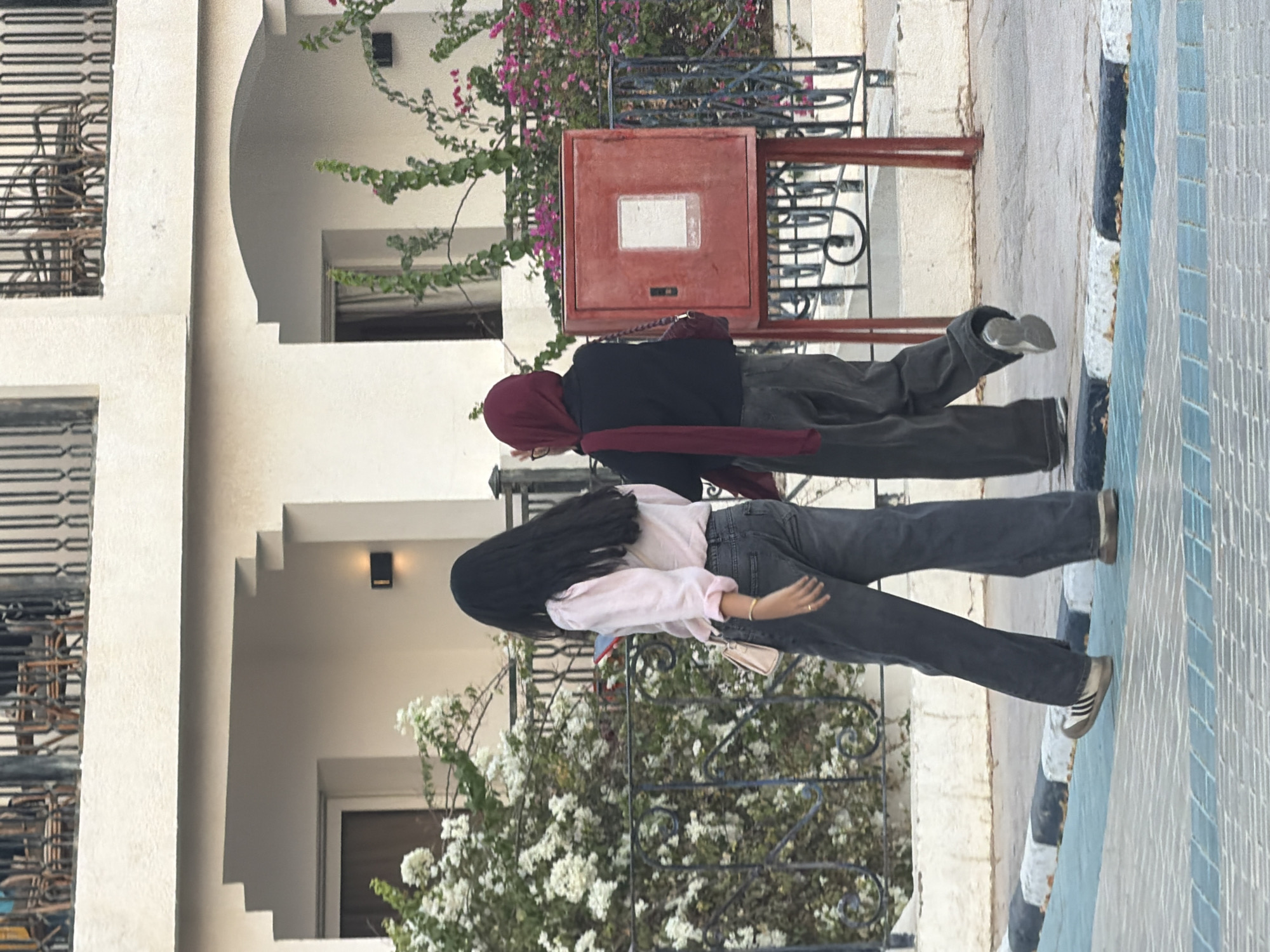 The image depicts two individuals walking away from the camera on a paved sidewalk. The person on the left has long, dark hair and is wearing a light pink long-sleeved shirt and dark grey flared jeans. They are carrying a small, light-colored purse and a mobile phone. The person on the right is wearing a dark top, a maroon hijab, and dark grey flared jeans. They are also carrying a dark-colored purse.
Behind them, a building with white stucco walls and arched doorways is visible. There are balconies with decorative railings on the upper floors, and flowering bushes with white and pink blooms are present along the building's facade and in front of a black metal fence. A red metal box, possibly a fire alarm or utility box, stands on a stand with black metal supports. The ground is paved with interlocking grey and blue tiles, bordered by a strip of black and white concrete curbing. The lighting suggests it is daytime. The scene is consistent with an outdoor environment in Sharm El Sheikh, Egypt, likely a resort area or public walkway.