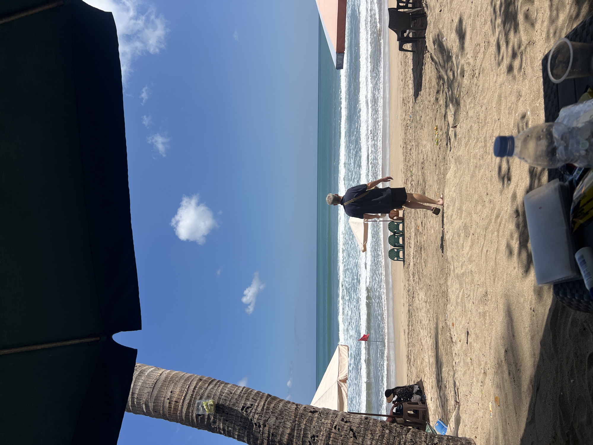 The image depicts a sunny day at Kuta Beach, Indonesia, with a clear blue sky and gentle waves rolling onto the sandy shore. In the foreground, a person is seen walking away from the camera towards the ocean, dressed in a dark blue polo shirt and black shorts. To the left, a large palm tree trunk is visible, with a small plastic cup containing some green material attached to it. Beach umbrellas, some light beige and one with orange and white stripes, are scattered across the sand. Several green plastic beach chairs are set up near the water's edge, and a red flag is planted in the sand. In the immediate foreground, a table holds a plastic water bottle, a white container, and a clear plastic cup. The scene conveys a relaxed atmosphere of a typical beach day.