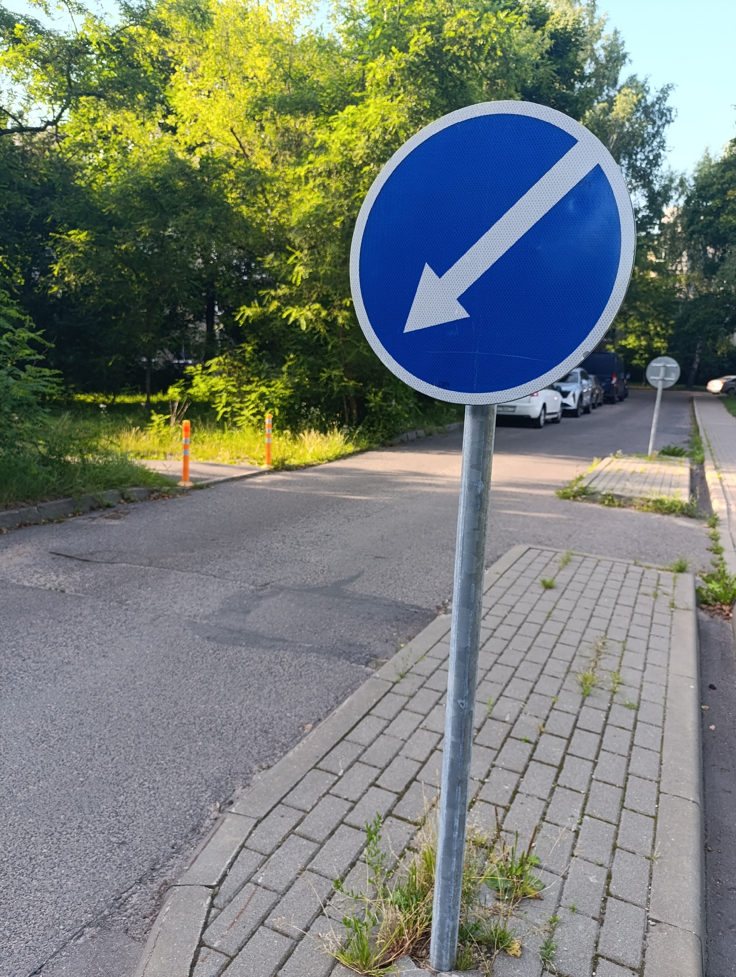 The image shows a blue circular traffic sign with a white diagonal arrow pointing down and to the left. The sign is mounted on a grey metal pole on the right side of a paved road. To the left of the road is a paved sidewalk with a curb. Green trees and foliage line the background, with a clear sky visible above. In the distance, several cars are parked along the side of the road. The scene appears to be a typical urban or suburban street in Minsk, Belarus, given the characteristic design of the traffic sign and the overall environment.