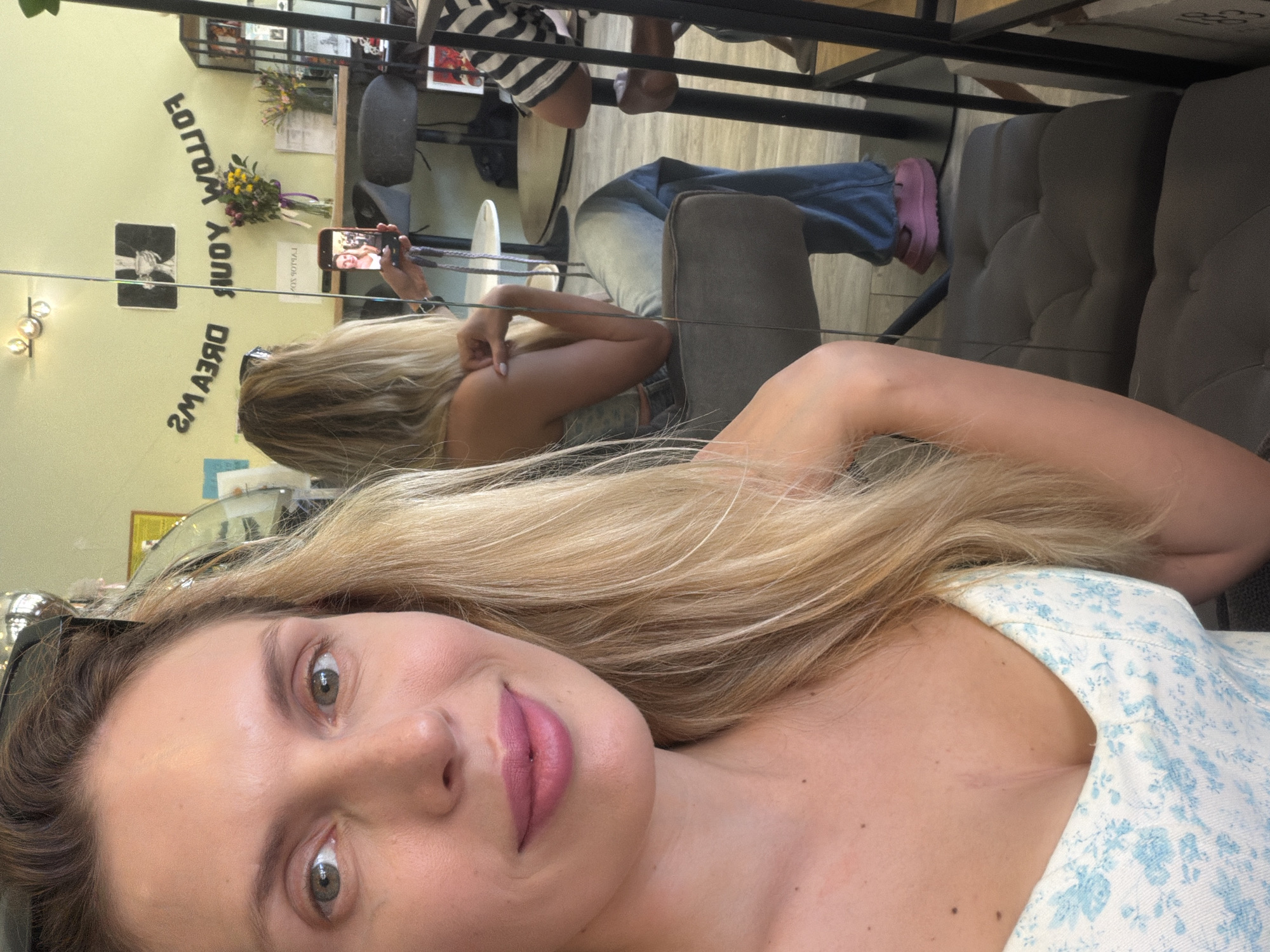 A blonde woman with light eyes and fair skin takes a mirror selfie inside what appears to be a cafe or lounge in Budva, Montenegro. In the foreground, the woman faces the camera directly, displaying a slight smile and wearing pink lipstick. She is dressed in a light-colored, blue floral pattern top and has dark sunglasses resting on her head.
The background reveals the woman's reflection in a mirror, where she is seated with her back partially turned, looking at a red smartphone held in her hands. The phone screen displays her own image, indicating the mirror selfie. She is wearing blue jeans and light pink slip-on shoes. She sits on a grey upholstered chair.
Behind the reflected woman, a light green wall features black, inverted block letters arranged in an arc, spelling "FOLLOW YOUR DREAMS." Above the text, a dark-framed picture or poster depicting a white trophy-like object is visible. Two small, spherical light fixtures are mounted on the wall above the text. To the right of the reflected woman, a wooden counter holds two vases containing colorful flowers. A person wearing a black and white striped shirt is partially visible seated at a table further to the right in the reflection. Additional tables, chairs, and a black shelving unit are discernible in the deeper background of the reflection.