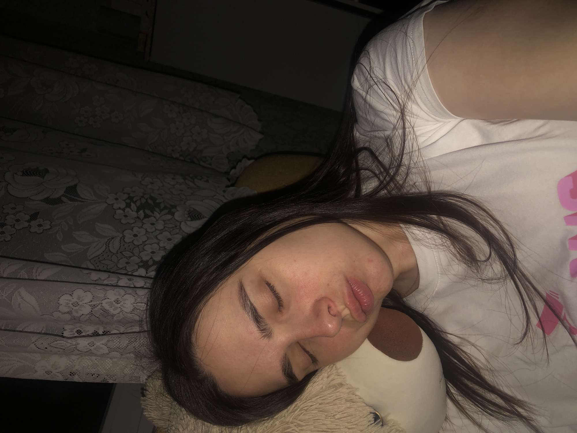 A young woman with long, dark hair is sleeping on a bed. She is wearing a white t-shirt with a pink graphic on it. Her head is resting on a beige and brown stuffed animal. The bed has a lace-patterned bedspread. The environment is dimly lit. There is no evidence that the image was taken in the city of Noviy, Russia.