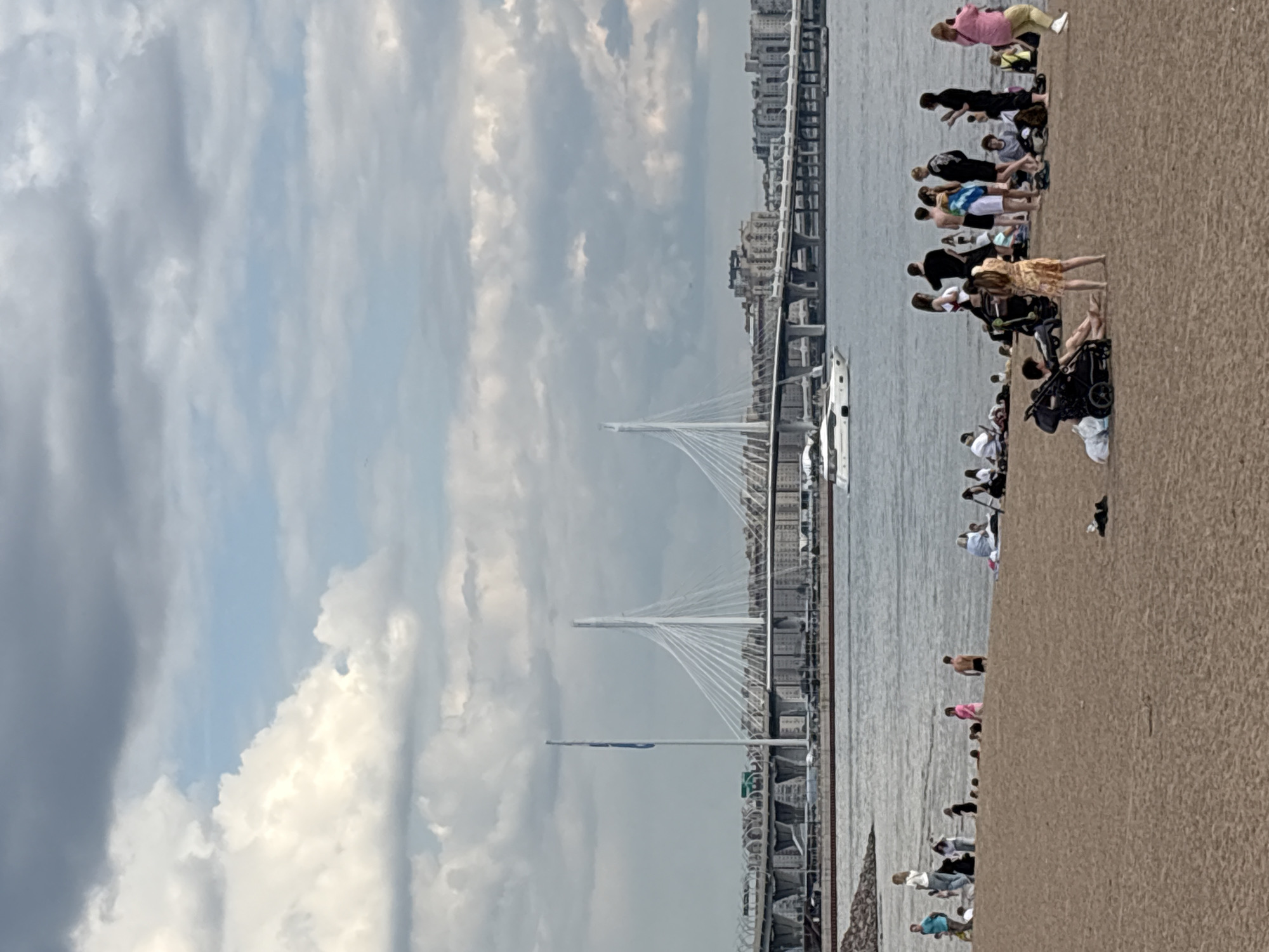This media file captures a lively daytime scene along a waterfront, likely in Saint Petersburg, Russia, based on the context. In the foreground, a wide, textured expanse, appearing as a sandy or paved promenade, is populated by numerous individuals. People of various ages are casually dressed, some in shorts and t-shirts, suggesting warm weather. Many are standing or walking, with some gathered closer to the water's edge, seemingly enjoying the view or the company. Several baby strollers are visible among the crowd, indicating families are present.
The middle ground features a large body of water, possibly an estuary or bay, upon which a white motor yacht with a dark roof is seen cruising, leaving a faint wake. Further into the middle ground, a tall flagpole with a blue flag is visible.
Dominating the background is a prominent modern cable-stayed bridge with multiple stark white pylons and a fan-like arrangement of numerous white cables supporting its deck. This architectural feature strongly suggests the Western High-Speed Diameter (WHSD) bridges in Saint Petersburg. Beyond the bridge, a city skyline with various buildings, some appearing tall and contemporary, stretches along the horizon.
The sky is partly cloudy, with a mix of bright white cumulus clouds and darker grey clouds, indicating a dynamic weather pattern but generally good visibility. The overall atmosphere appears leisurely and recreational, with people enjoying an outdoor setting by the water. No discernible text is visible within the image.
