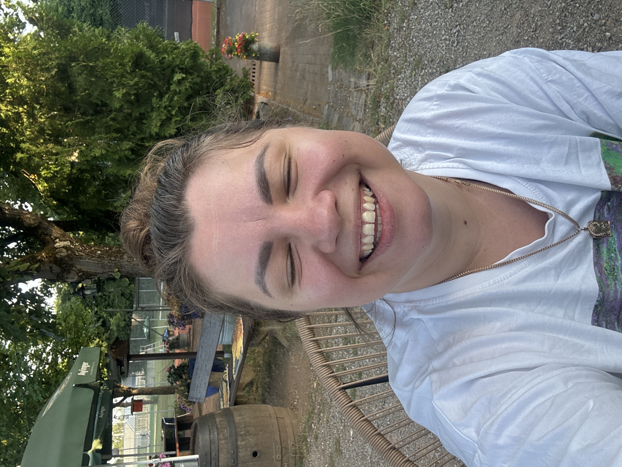A woman with her eyes closed and a wide smile is taking a selfie outdoors. She has her hair pulled back and is wearing a white t-shirt with a gold chain necklace. Behind her, a tree with green foliage is visible, along with a gravel path and a wicker chair. In the background, there are glimpses of what appears to be a courtyard or outdoor seating area with a barrel and some flowers in planters. The lighting suggests it is daytime, possibly late afternoon, with the sun casting shadows. The setting appears to be casual and relaxed. There is no visible text in the image.