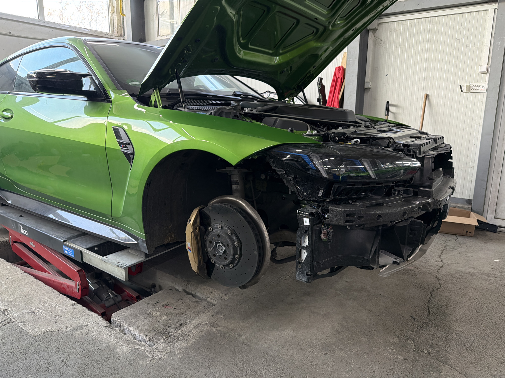 The image depicts a green BMW M4 coupe with its hood open and the front bumper removed, revealing its internal components. The car is elevated on a hydraulic lift in what appears to be a repair shop or garage. The front left wheel is also removed, exposing the brake rotor and caliper. The setting is an indoor space with a concrete floor and white walls. Based on prior knowledge, the location is identified as a service shop in Burgas, Bulgaria. There are no visible people in the image. The environment suggests a work in progress, focusing on the mechanical aspects of the vehicle.