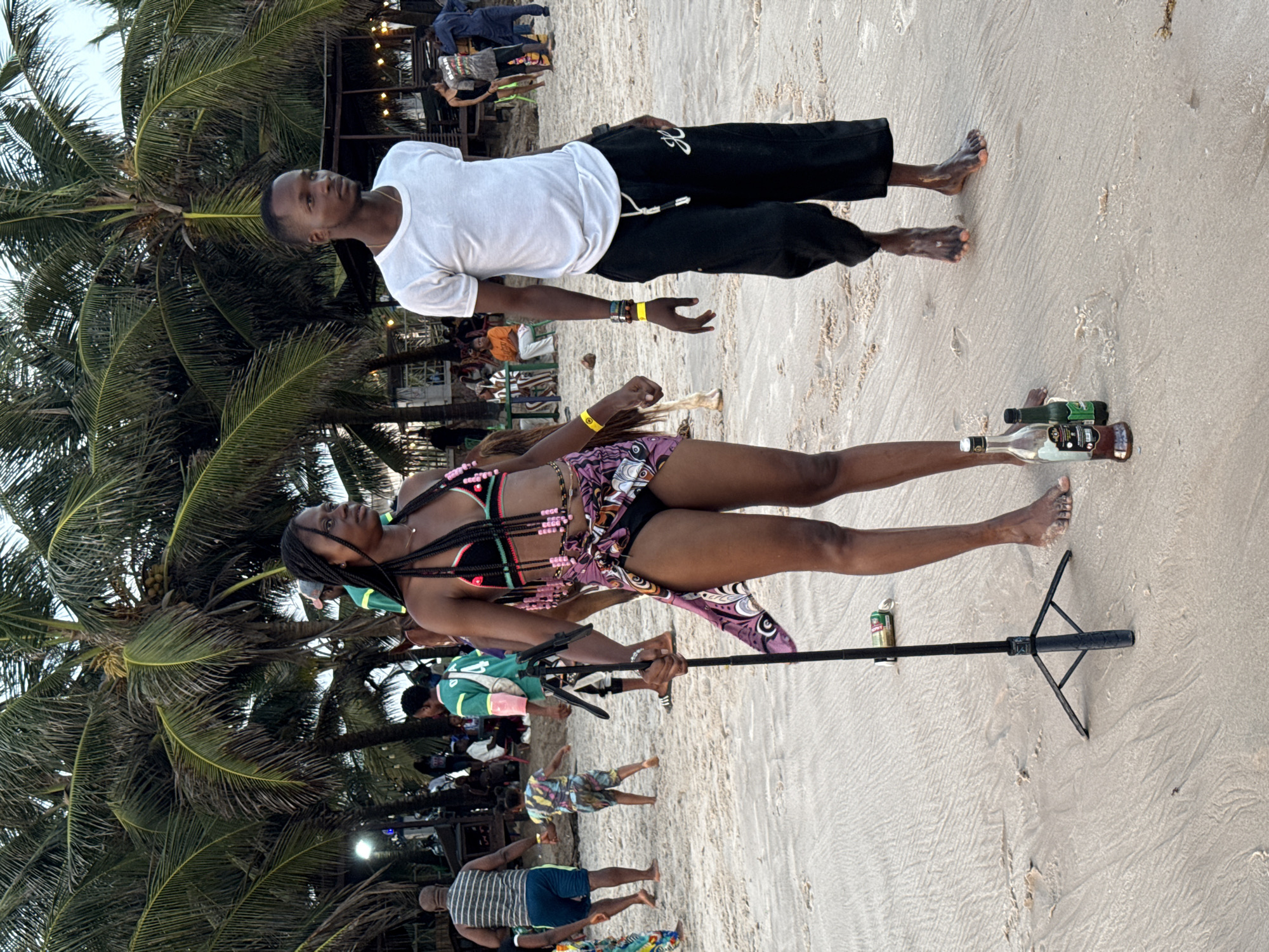 A beach scene in Mopo Onijebu, Nigeria, features two individuals prominently. A man in a white t-shirt and black pants stands near a woman in a bikini top and sarong. She holds a black selfie stick. Two bottles and a can of beverage sit on the sand near the selfie stick. Several other people are in the background, and palm trees are visible.