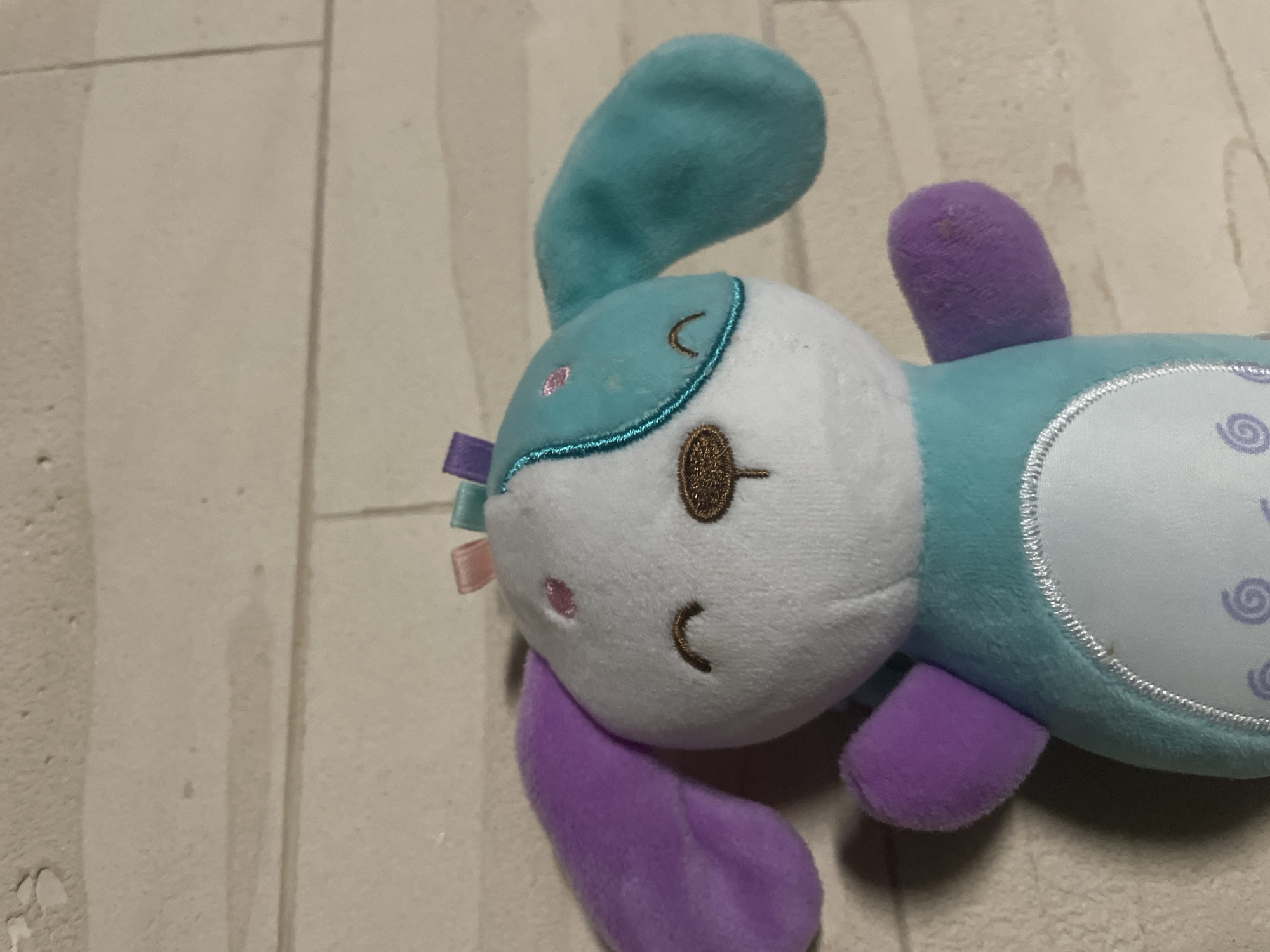 A plush, possibly toy, bunny or similar animal lies on a surface that appears to be light-colored wood grain laminate flooring. The toy has a predominantly white face with a teal-colored patch on top. It features embroidered facial features. The ears are teal and purple, and the body is teal with white sections featuring a swirl pattern. Several ribbons are attached to the side of the head. The image provides no information as to the location of Novi Sad, Serbia.