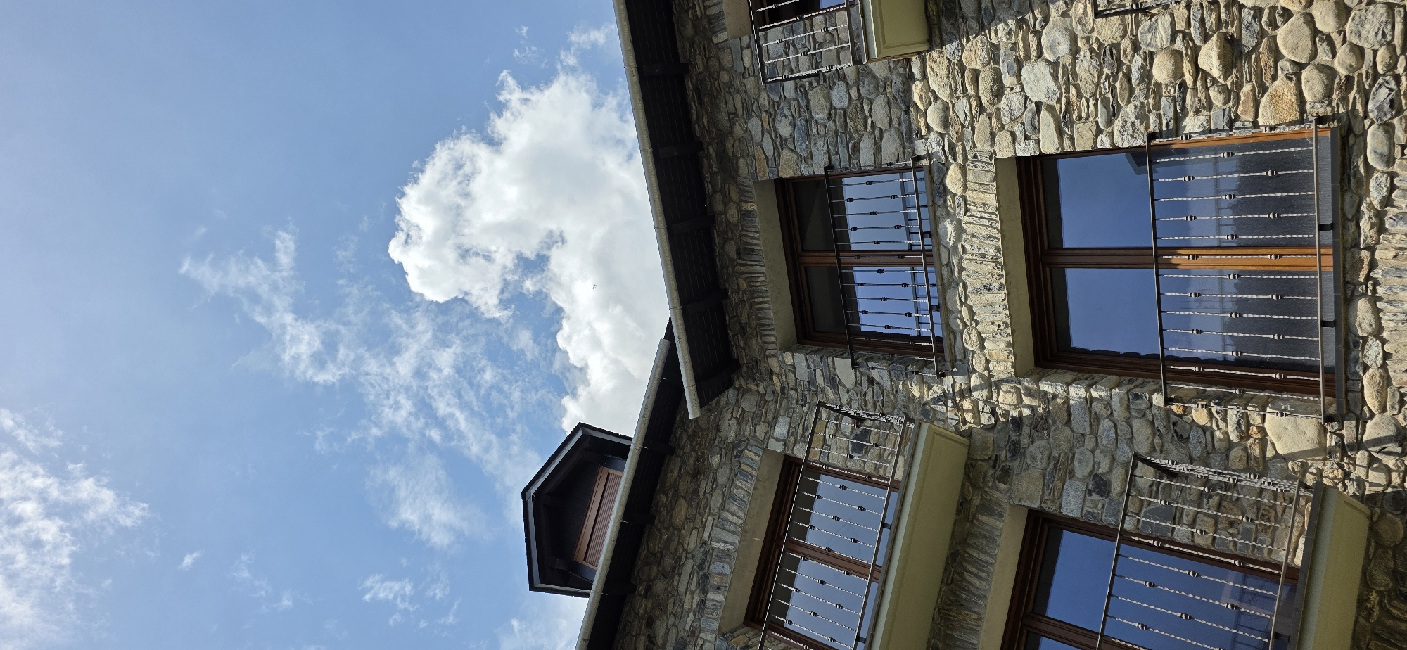 The image presents an upward view of a stone building set against a bright blue sky with scattered white clouds. The building's exterior is constructed of varying sizes and colors of stones. It includes windows with dark wood frames and decorative wrought iron bars, reflecting the sky. The perspective is angled, tilting the building diagonally across the frame. This image was probably taken in the city of Benasque, Spain.