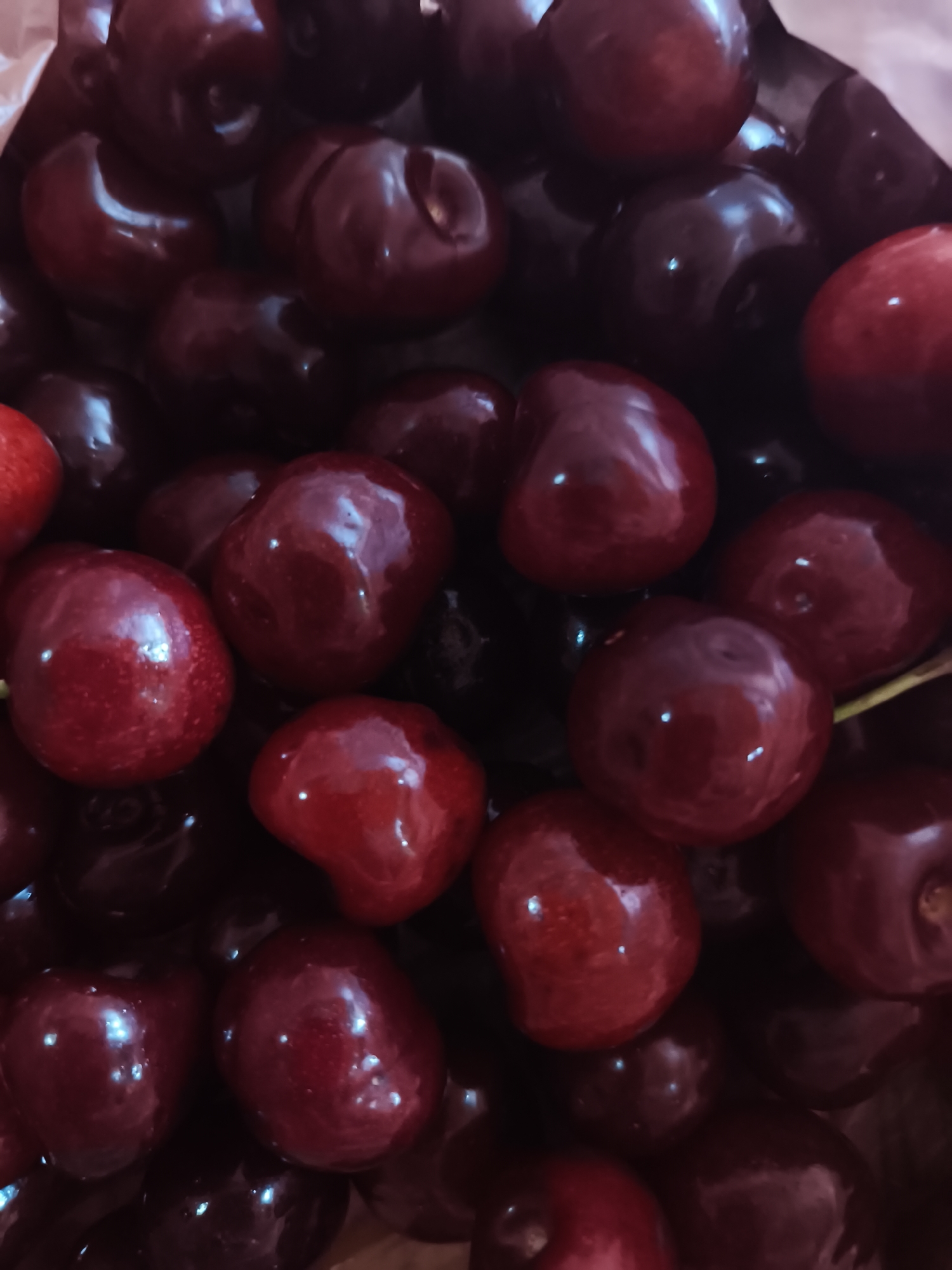 The image is a close-up, top-down shot of a pile of dark red cherries. The cherries are plump and glossy, with some reflecting bright white highlights from the light source. The background is dark, obscuring any details of the environment. There are no people or other objects visible in the image. The location is unknown.