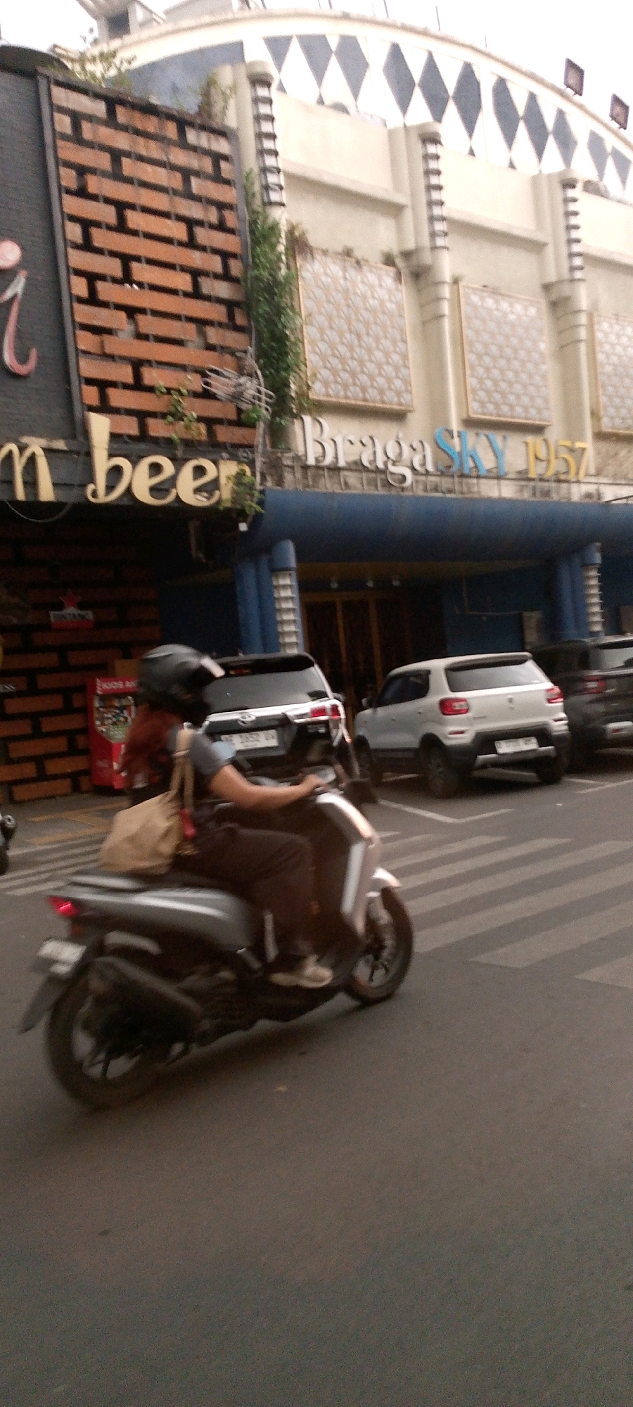 A motorcycle rider, wearing a helmet and a light brown bag slung over their shoulder, is moving left to right in the frame on a street with a crosswalk. The rider is positioned on a silver scooter, which is the primary subject. In the background, there are parked cars, including a white SUV and a black SUV. Behind the vehicles, a building is visible with signage that reads "Braga SKY 1957" and "m beer". The architecture of the building features brickwork and decorative panels. The scene appears to be a street in Bandung City, Indonesia, given the context of the signage.
