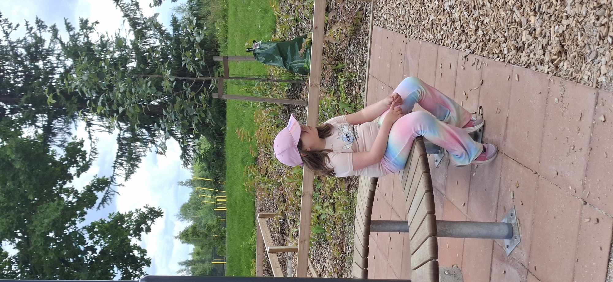 A young girl, wearing a pink baseball cap, a light pink t-shirt with an animated character on it, and colorful tie-dye pants in shades of pink, blue, and yellow, sits on a wooden slatted bench. She is facing to her right, looking away from the camera. Her hands are resting in her lap. The bench has metal supports and is situated on a paved area with reddish-brown square tiles. Scattered wood chips are visible on the ground around the bench and to the right of the paved area.
In the background, a wooden fence encloses an area with small plants. Beyond this, a grassy field stretches out, with several tall, thin yellow poles visible in the distance, possibly part of a playground or sports facility. A large evergreen tree stands behind the girl, with a smaller sapling nearby protected by stakes and a green bag. Other trees and foliage are visible further back, and the sky is overcast with some clouds. The overall scene appears to be an outdoor public space, likely a park or recreational area, located in Lahti, Finland.