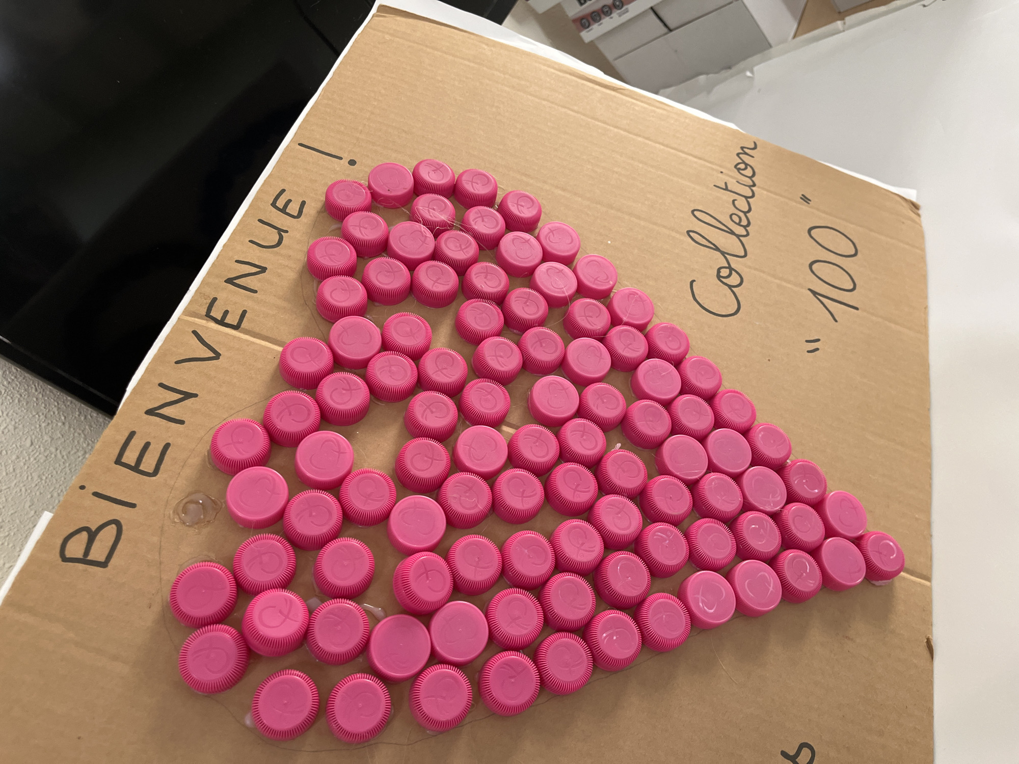A slightly high-angle indoor shot depicts a brown cardboard box with handwriting and pink bottle caps arranged in a heart shape. The words "BIENVENUE!" are written on the left side of the box. On the upper right, "Collection" and "100" are written. The pink bottle caps, each with a white logo, form a heart shape, glued to the cardboard. A flat-screen television can be partially seen in the upper left corner, while what appears to be a white desk or shelf is on the right. The location is the city of La Fouillouse, France.