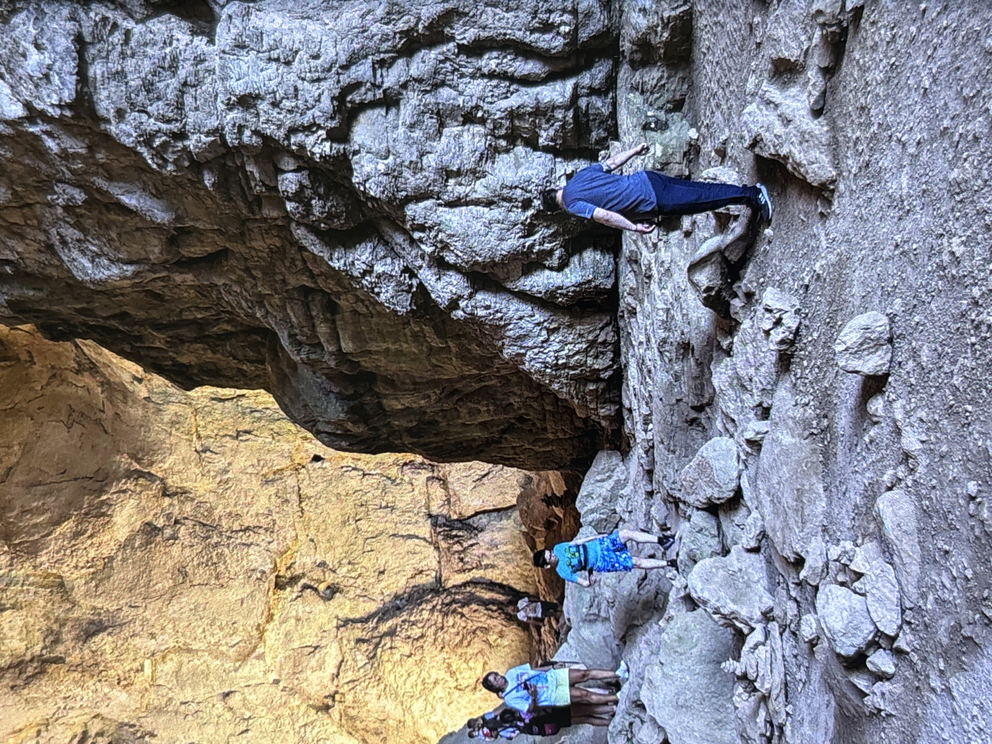 The image depicts a scene inside a large, rocky cavern or deep gorge. The perspective suggests looking upwards at towering rock formations. On the right side of the frame, a person wearing a blue t-shirt and dark trousers is actively climbing or traversing a very steep, textured grey rock face. Their body is angled significantly, with arms extended and feet finding purchase on small ledges, indicating a challenging ascent or descent.
Below this climber, on a lower rocky path or ledge, four other individuals are visible. One person wears a blue patterned t-shirt and blue shorts, while another, positioned slightly further back, wears a light-colored t-shirt and dark shorts, holding what appears to be a bag. Two other people are partially visible behind them. All individuals appear to be engaged in exploration or hiking within this natural environment.
The rock formations vary in color and texture; the upper and right portions of the cave are composed of dark grey, highly textured rock, while the lower-left section is a lighter, yellowish-brown hue, possibly illuminated by a natural light source filtering in or a different type of rock. The setting evokes a sense of adventure and natural grandeur. There is no visible text in the image. Given the provided context, the scene is within a natural cave or gorge, possibly in the region of Wolverhampton, United Kingdom, if such a formation exists there.