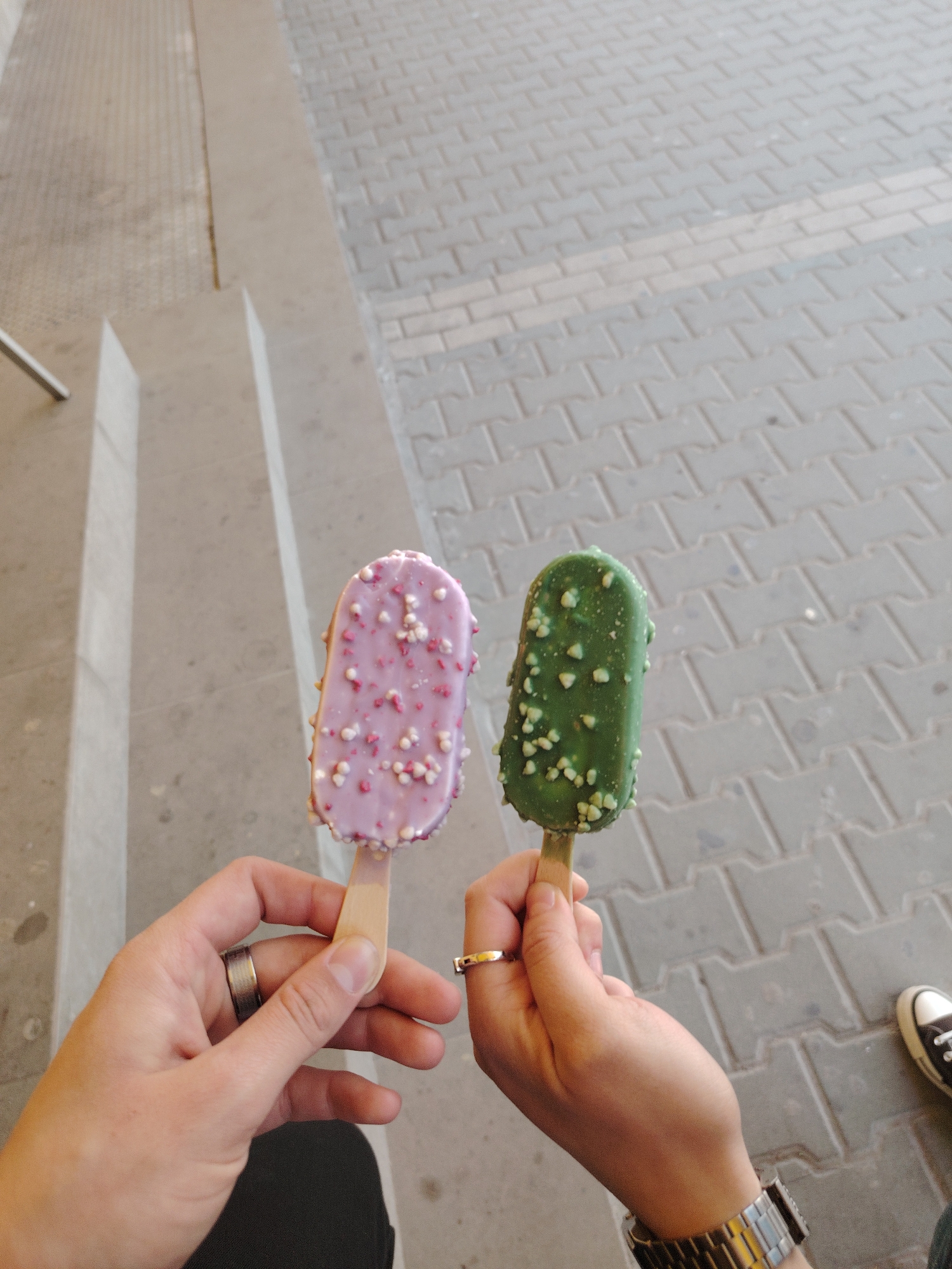 Two hands hold two ice cream bars against an outdoor backdrop. The hand on the left holds a pink or light purple ice cream bar adorned with white and pink sprinkles. This hand wears a broad, silver-toned ring on its ring finger. The hand on the right holds a green ice cream bar, which is covered with light green or white chunky sprinkles. This hand has a thin gold-colored ring on its ring finger and a silver-toned watch on its wrist.
The setting appears to be a public outdoor space during the daytime. On the left side of the frame, there is a grey concrete structure, possibly stairs or a ledge, with a sliver of a metal railing visible. The ground consists of light grey interlocking pavers, typical of urban walkways. In the bottom right corner, a partial view of a black canvas shoe with a white sole is visible. There is no visible text in the image. The scene depicts a casual moment of enjoying frozen treats.