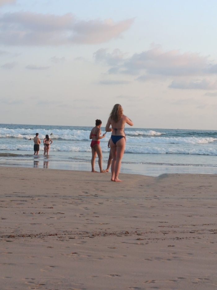 The image depicts a beach scene at Kabalana, Sri Lanka, under a partly cloudy, light blue sky with hints of pink. The foreground consists of damp, light brown sand with visible footprints. In the mid-ground, five individuals are present. On the dry sand, two females stand with their backs mostly to the viewer: one on the right, with light, wavy hair, wears a dark blue two-piece swimsuit and faces the ocean; slightly to her left and behind, another female with dark hair wears a red two-piece swimsuit and faces partially right, towards the ocean. Further left, in the shallow ocean water where small waves are breaking, a male in dark shorts and a female in a dark two-piece swimsuit stand, both facing the ocean. A fifth individual is partially visible further out in the ocean amidst the waves, possibly with a surfboard. The ocean exhibits multiple breaking waves. No objects other than sand, water, and clothing are explicitly visible in detail.