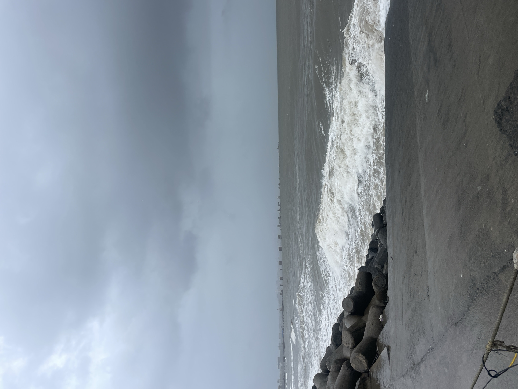 The image depicts a scene at Mumbai's Bandra Worli Sea Link, captured during a cloudy or potentially stormy day. In the foreground, large concrete tetrapods are arranged on a gray, paved surface, likely a walkway or breakwater. These structures are designed to dissipate wave energy.
Beyond the tetrapods, the choppy, gray sea is visible, with whitecaps indicating moderate to rough conditions. Waves are crashing against the shore. In the distance, across the sea, the Mumbai skyline is faintly visible, characterized by buildings and what appear to be construction sites or incomplete structures. The silhouette of the Bandra Worli Sea Link's pylons and cables can be discerned stretching across the horizon.
The sky is overcast with heavy, dark gray clouds, suggesting the possibility of rain or an impending storm. The overall atmosphere is somber and dramatic, reflecting the turbulent weather. There are no people visible in the frame, and the focus is on the natural elements and the man-made infrastructure. The image seems to have been taken from an elevated position looking out towards the sea and the distant cityscape.