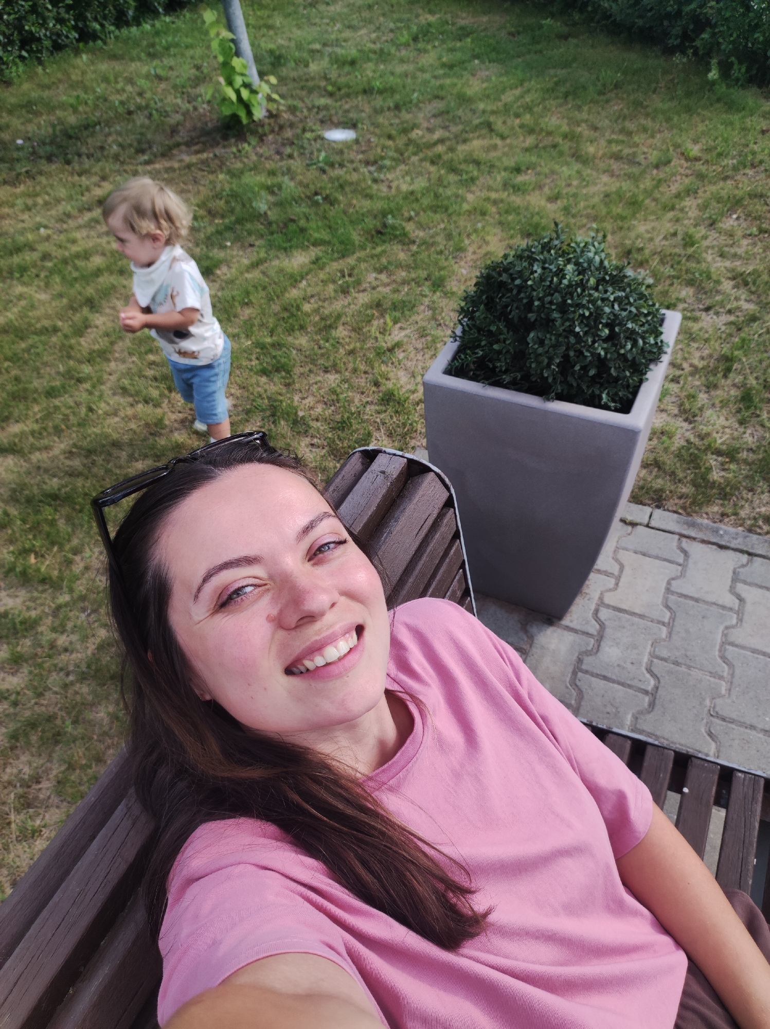 A woman with long, dark hair and a pink t-shirt smiles at the camera, holding the camera for a selfie. She is sitting on a wooden bench outdoors. In the background, a young child with blond hair, wearing a white t-shirt and blue shorts, stands on a grassy area. A grey planter with a green shrub sits to the right of the bench, and paving stones are visible on the ground. The scene appears to be a park or a backyard in Chaikovsky, Russia.