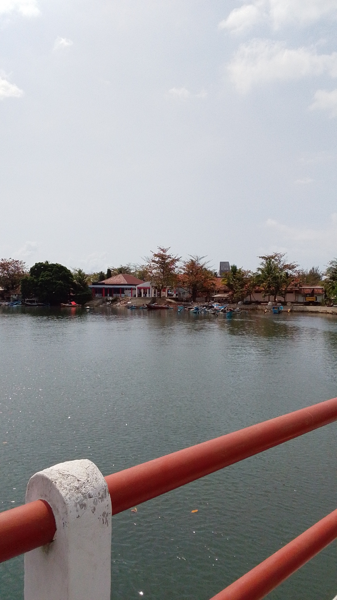 A wide, static, eye-level outdoor shot captures a tranquil scene in Desa Karangjaladri, Indonesia, dominated by a calm body of water. In the foreground, a weathered red railing and a white concrete support post of a bridge or walkway frame the lower portion of the image, hinting at the vantage point from which the photo was taken. Beyond the railing, the water stretches towards the horizon, reflecting the bright, overcast sky and showing small glints of sunlight.
Along the far bank, a cluster of buildings and trees creates a serene shoreline. A prominent building with a red-tiled roof, featuring white pillars and red trim, stands out. Several smaller structures with similar roofing are visible to its left and right. A variety of trees, some with reddish-brown foliage, punctuate the landscape, including a large, dark green tree on the left side. Small boats, mostly light-colored and some appearing to be fishing vessels, are moored or gently floating near the shore, suggesting a coastal or riverine community. In the far distance, a tall, slender structure, possibly a tower or lighthouse, is visible against the sky. The overall impression is one of peaceful, everyday life in a waterside village.