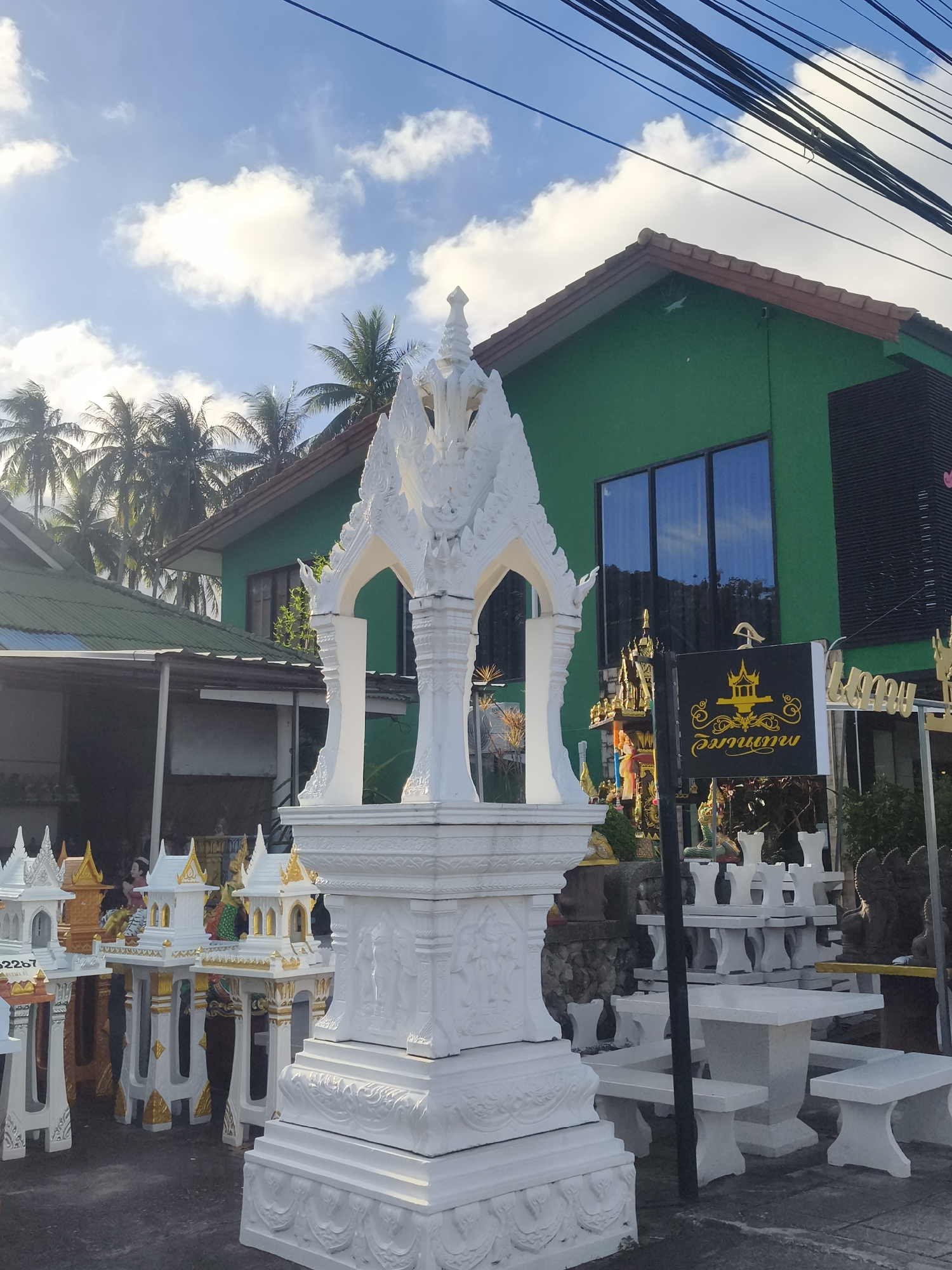 A collection of small, ornate spirit houses and a large white shrine are displayed outdoors in front of a bright green building. The spirit houses are intricately designed, many with white walls and gold accents, featuring tiered roofs and decorative pillars. The large central shrine is also white, with elaborate carvings and a multi-tiered roof structure. In the background, the green building has large windows reflecting the sky and palm trees. A sign with yellow Thai script on a black background is visible. The scene appears to be set in a shop or marketplace specializing in these religious structures, likely in Baan Thong Po, Thailand. Palm trees are visible, suggesting a tropical location. The sky is blue with scattered white clouds, and the lighting indicates it is daytime, possibly morning or afternoon. There are no people clearly visible in the image, but the offerings and decorations suggest a place of worship or commerce related to spiritual practices.