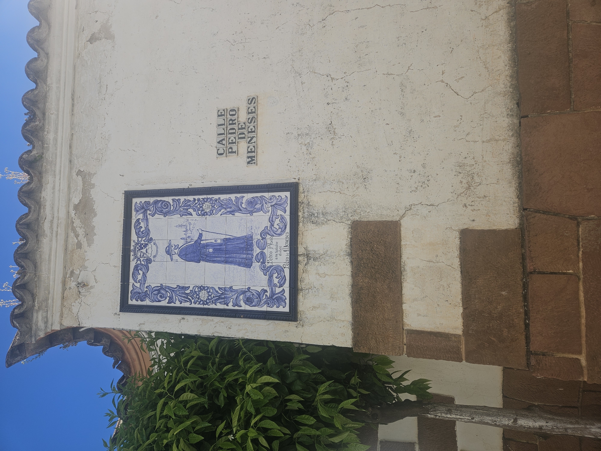 The image shows a building corner in Pedro Abad, Spain, marked by a street sign that reads "Calle Pedro de Meneses" (Pedro de Meneses Street). A blue and white tiled plaque depicting a figure, likely related to the street's namesake, is affixed to the white wall. The wall has cracks and an aged appearance. Below the plaque, green foliage partially obscures the base of the wall. The building features a scalloped edge detail and brickwork accents. The sky is visible in the background.