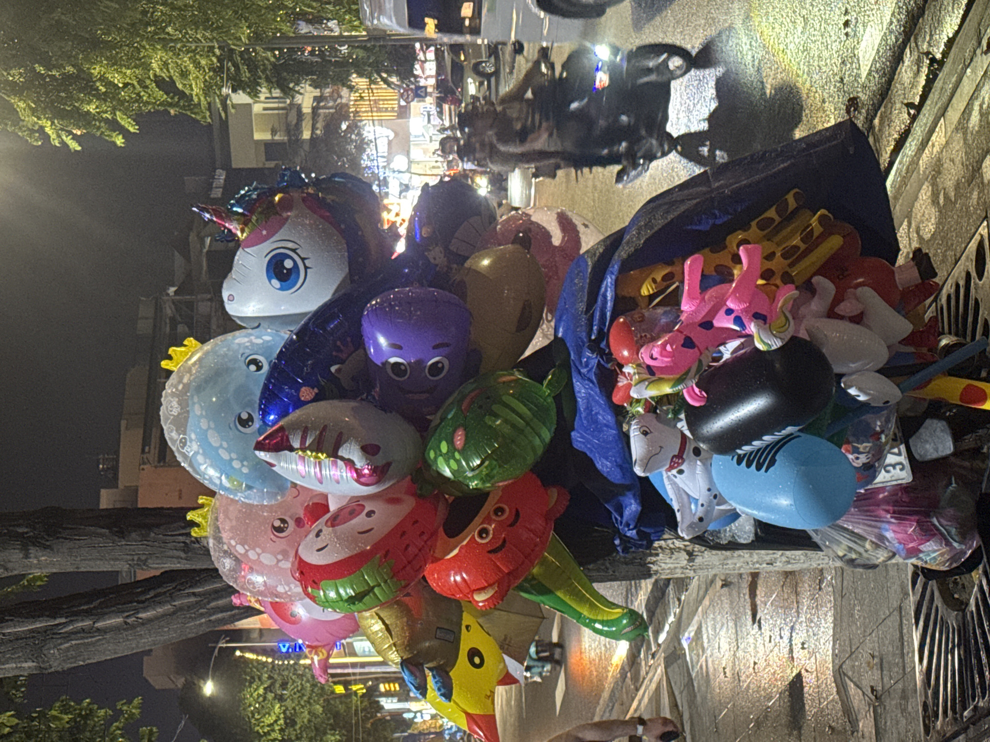The image shows a vendor's balloon and toy stand in Nha Trang Ward, Vietnam, at night. The stand, covered with a blue tarp, displays an assortment of colorful, inflatable balloons shaped like animals and cartoon characters, including a unicorn, octopus, and watermelon. Stuffed toys, including giraffes and dogs, are also visible. The scene is on a city street, with reflections indicating wet pavement. Streetlights and vehicle lights illuminate the background. Motorbikes and other vehicles are visible in the street behind the stand.