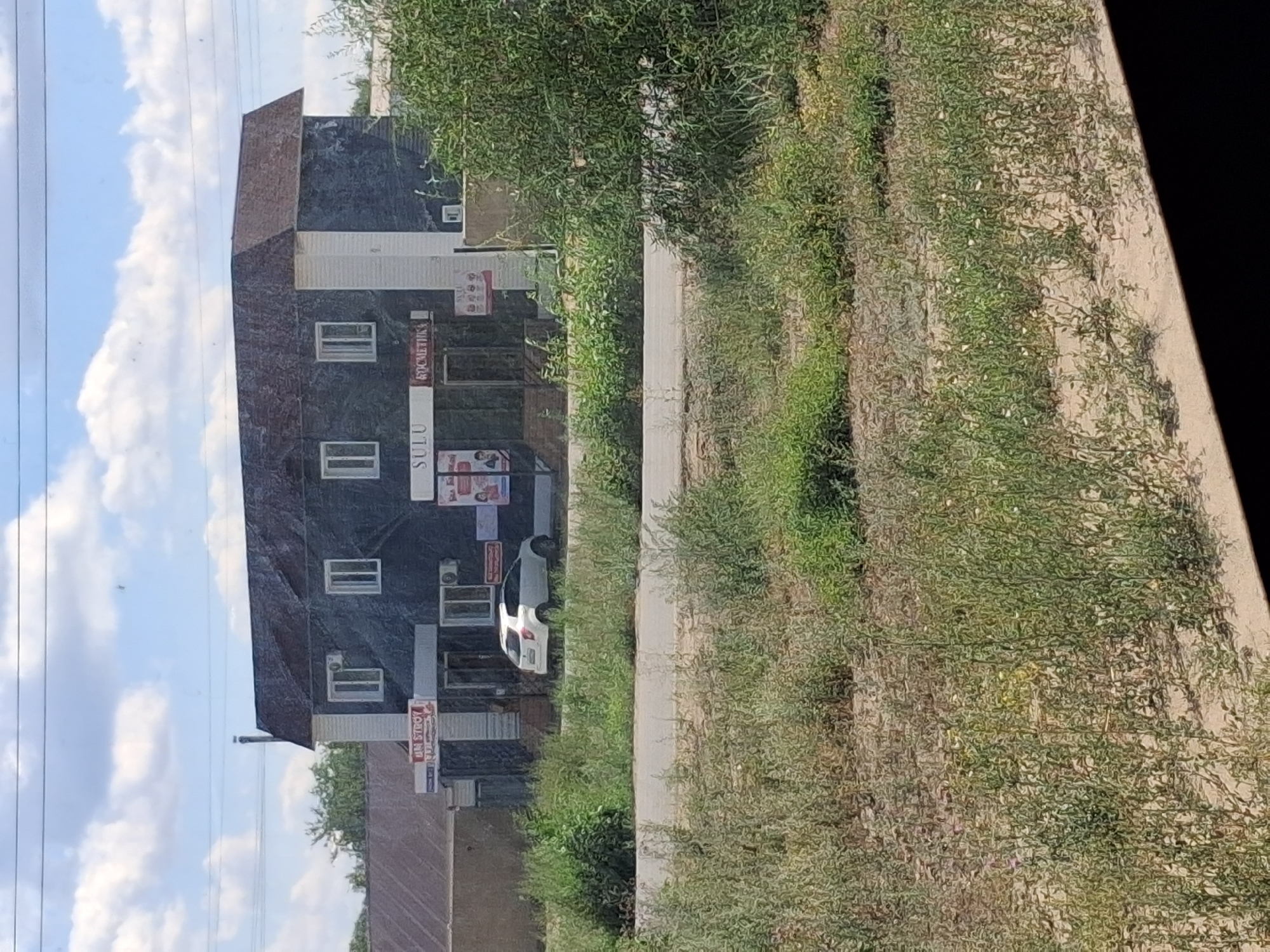 The image captures a street-level view of a two-story building with a dark, textured facade and white architectural accents, situated in Semey, Kazakhstan. The building houses several businesses, indicated by signage. To the left, a sign reads "UM STROY," suggesting a construction-related business. In the center, a prominent sign displays "SULU," and to its right, another sign reads "КОСМЕТИКА," indicating a cosmetics store. A white hatchback car is parked in front of the building, visible through the foreground foliage and a paved road. The environment is rural or semi-rural, characterized by overgrown vegetation and a clear sky with scattered clouds. The overall scene depicts a commercial area with moderate business activity.