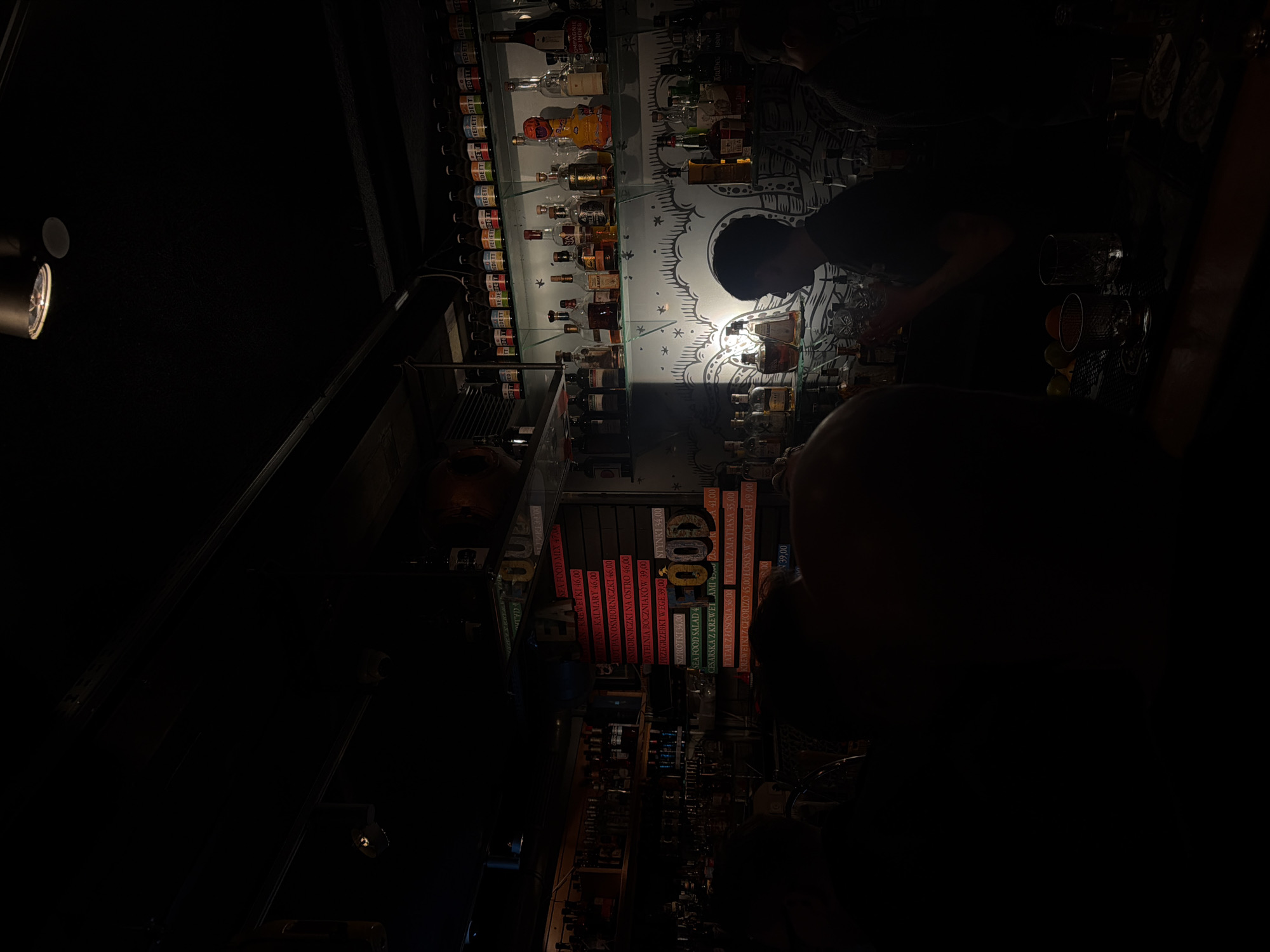 The image depicts the interior of a dimly lit bar. A bartender is visible behind the counter, preparing drinks amidst numerous bottles of liquor and glassware. A food menu displays dishes priced in Polish currency, possibly indicating the location is in Poland. Patrons can be discerned in the foreground, though they are mostly obscured by shadow. The atmosphere is casual and intimate due to the low lighting.