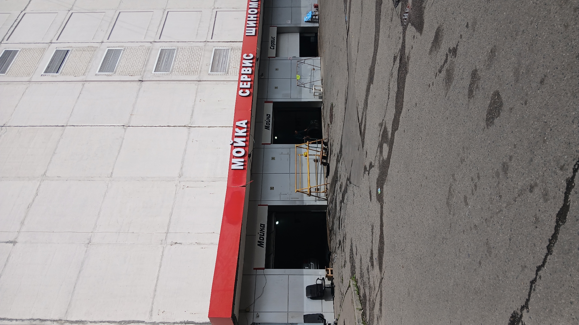 A street-level, exterior, full-shot of a car wash and service center with a prominent red awning. The building has a white concrete facade with horizontal and vertical paneling. The awning has large white Russian letters that spell out "МОЙКА СЕРВИС ШИНОМОНТАЖ" (Car wash Service Tire fitting). Below the awning, there are several bays, two of which are open, displaying signs for "Мойка" (Car wash) and "Сервис" (Service). One bay on the left has a car inside, and another bay on the right has a person working on a car. Yellow metal scaffolding is set up in front of one of the bays. The ground in front of the building is asphalt, with some puddles and cracks. Two black office chairs are positioned to the left of the first bay. The scene is illuminated by daylight.