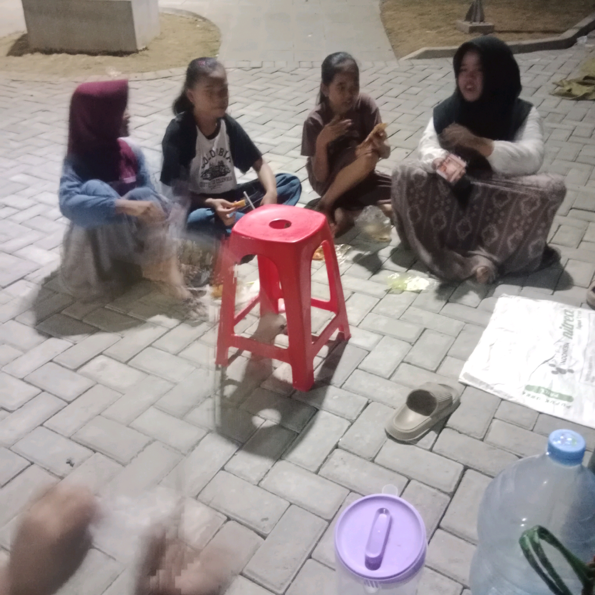 In Tlogorejo, Indonesia, four individuals, likely children or young adults, are seated on a paved area, possibly a park or public space, around a red plastic stool. The ground is covered with light gray interlocking bricks arranged in a herringbone pattern. One person on the far left wears a maroon hijab and a blue garment. To their right, another individual in a black and white t-shirt and blue pants holds a small white object. The third person, in a brown shirt, is holding a piece of food. The fourth individual, on the far right, is wearing a black hijab and a white top with a patterned skirt, and is holding some banknotes. Various objects are scattered around them, including a white bag labeled "NITREA", a pair of brown sandals, a purple lidded container, and a large clear plastic water jug with a blue cap. The lighting suggests it is evening or night.
