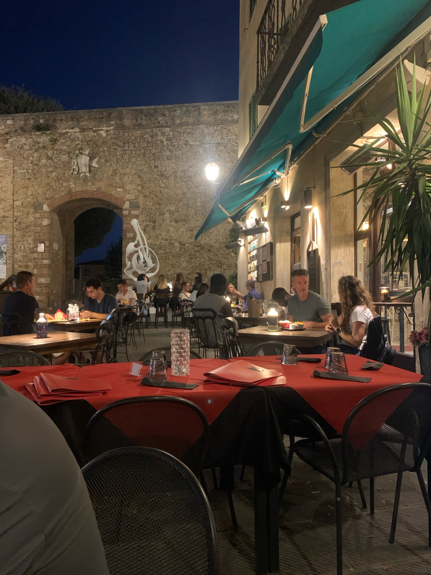 An exterior shot at dusk shows a busy outdoor dining area in San Quirico d'Orcia, Italy. Tables are set with red tablecloths and black chairs. Several patrons are seated, eating and conversing. The restaurant appears to be located next to an ancient stone wall with an arched gateway. The atmosphere is lively and convivial, with the warm glow of overhead lights illuminating the scene.