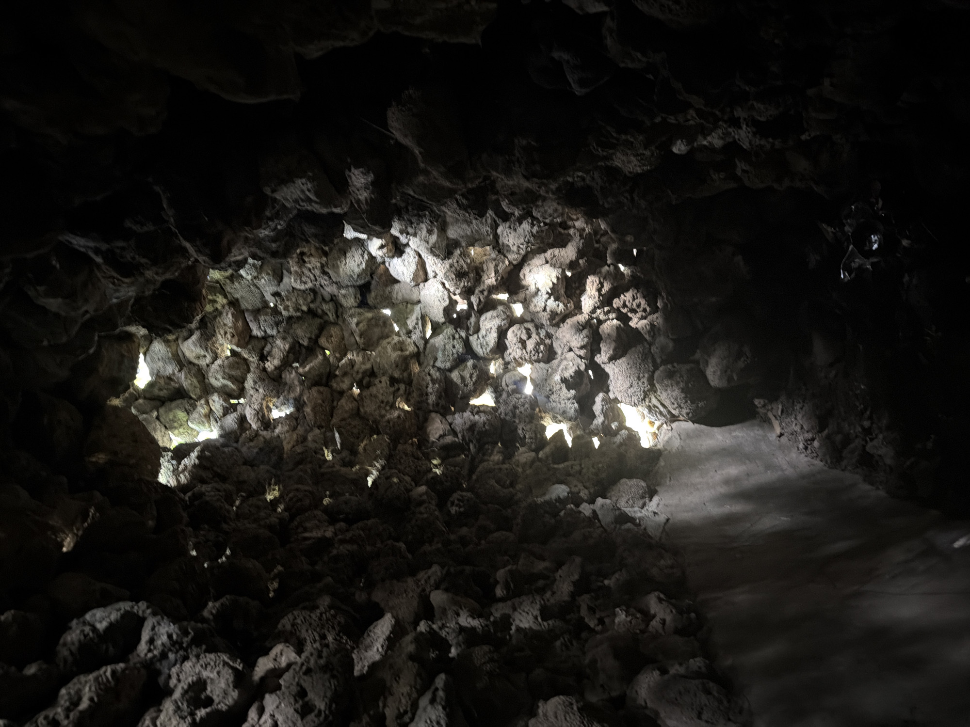 The image captures the interior of a dark, cavernous space, likely a cave or grotto, characterized by rough, dark grey rock formations. The scene is dominated by natural light filtering through numerous irregular openings between the rocks, primarily on the left and central parts of the frame. These bright shafts of light create dramatic contrasts, illuminating the rugged, porous texture of the stone walls and casting deep shadows in other areas.
The ceiling and walls are composed of densely packed, irregular stones, possibly volcanic rock, which create a raw and untamed atmosphere. No people are visible, and no specific activity is taking place. On the right side of the frame, a smoother, lighter-toned pathway or floor is discernible, suggesting a maintained area for visitors to traverse the dark environment. This pathway contrasts with the wilder, more chaotic rock formations. The strong directional light from outside indicates it is daytime. The overall impression is one of natural beauty and geological interest, with the interplay of light and shadow highlighting the cave's unique features. No text is visible in the image.
