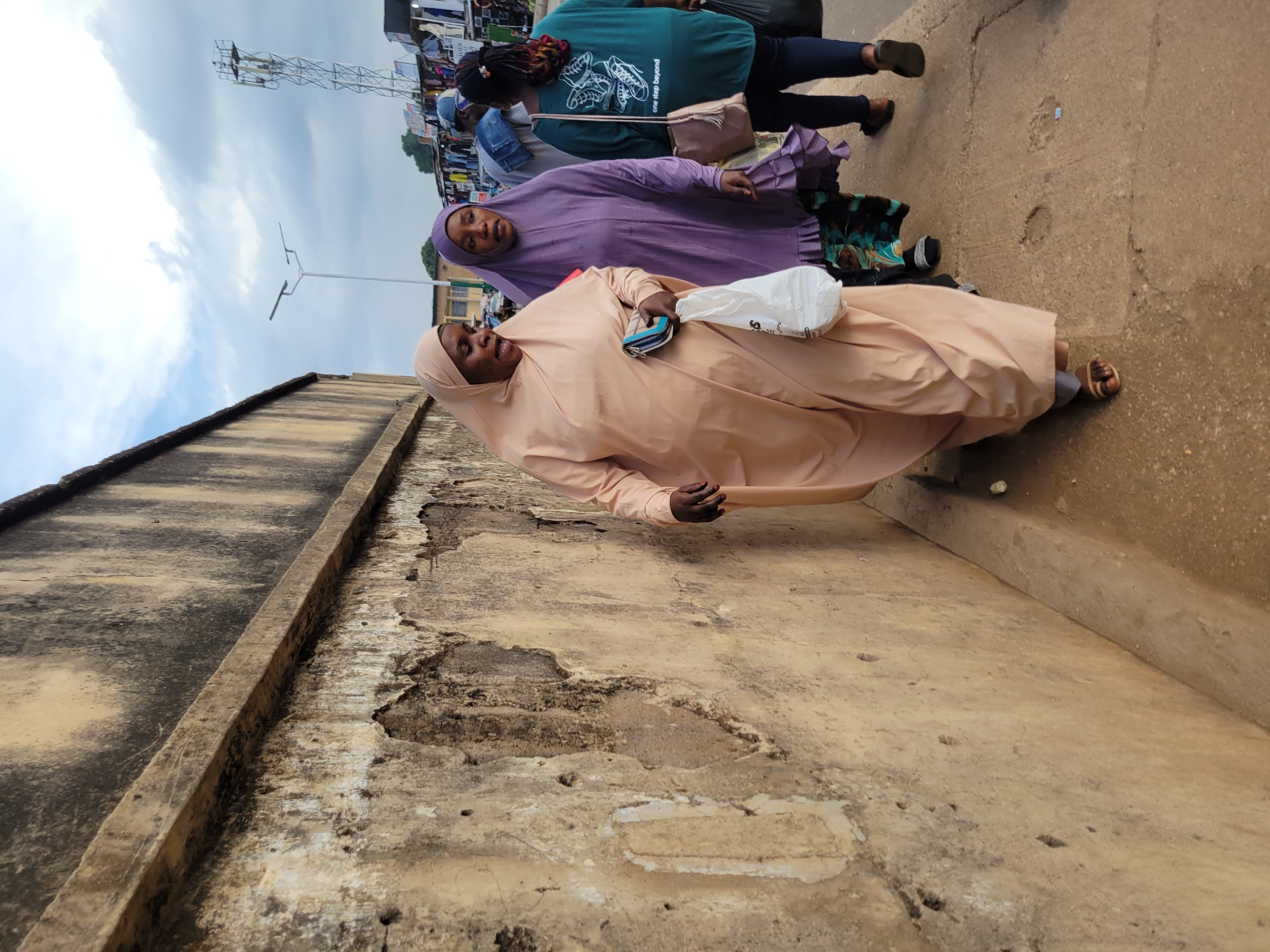 The image depicts a street scene in Jos, Nigeria, characterized by a weathered, textured concrete wall dominating the left foreground. The wall exhibits signs of age and decay, with patches of discoloration and crumbling surfaces. Three women are walking away from the viewer on a concrete walkway that runs alongside the wall.
The woman in the foreground on the left is wearing a light peach-colored hijab and abaya. She is carrying a white plastic bag in her left hand and a small wallet or clutch in her right hand. Her head is turned slightly to her right, and her mouth is open as if she is speaking.
Behind her, a woman in a vibrant purple hijab and matching abaya walks in the same direction. Her gaze is directed forward, and she appears to be looking ahead.
To the right of these two women, a third woman is partially visible. She is wearing a green t-shirt with a graphic design of shoes and text that reads "one step beyond." She is also carrying a bag over her right shoulder.
In the background, a bustling market scene unfolds. Stalls with various goods are visible, and other pedestrians can be seen. A tall communication tower stands prominently in the distance, alongside a street lamp. The sky is partly cloudy, with a mix of white clouds against a lighter blue sky. The overall impression is of a lively, everyday scene in an urban Nigerian setting.