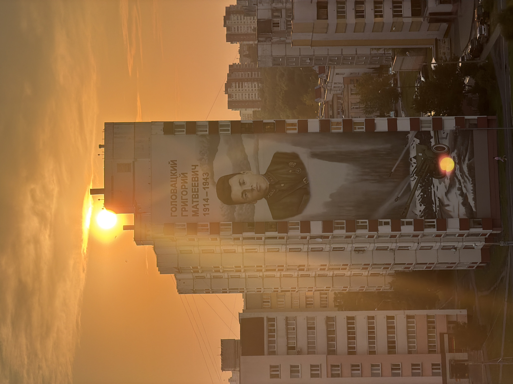 The image captures a mural on the side of a residential building in Homyel, Belarus, during sunset. The mural depicts a portrait of a man in military uniform, identified by Cyrillic text above his image as "ГОЛОВАЦКИЙ ГРИГОРИЙ МАТВЕЕВИЧ 1914-1943" (Golovatsky Grigory Matveevich 1914-1943). Below the portrait, a depiction of a military cannon firing is visible, with the setting sun appearing as a red orb over the cannon's barrel. The building itself is tall, multi-story, with numerous windows. The sky is filled with warm, orange hues characteristic of sunset, with clouds present. Several other apartment buildings of varying heights surround the main building, some in the foreground and others in the background, contributing to a dense urban landscape. Trees are visible between the buildings in the mid-ground and background. The sun's glare is strong, affecting the clarity of some parts of the image. The overall scene suggests a memorial dedicated to a war hero in a residential area of the city.