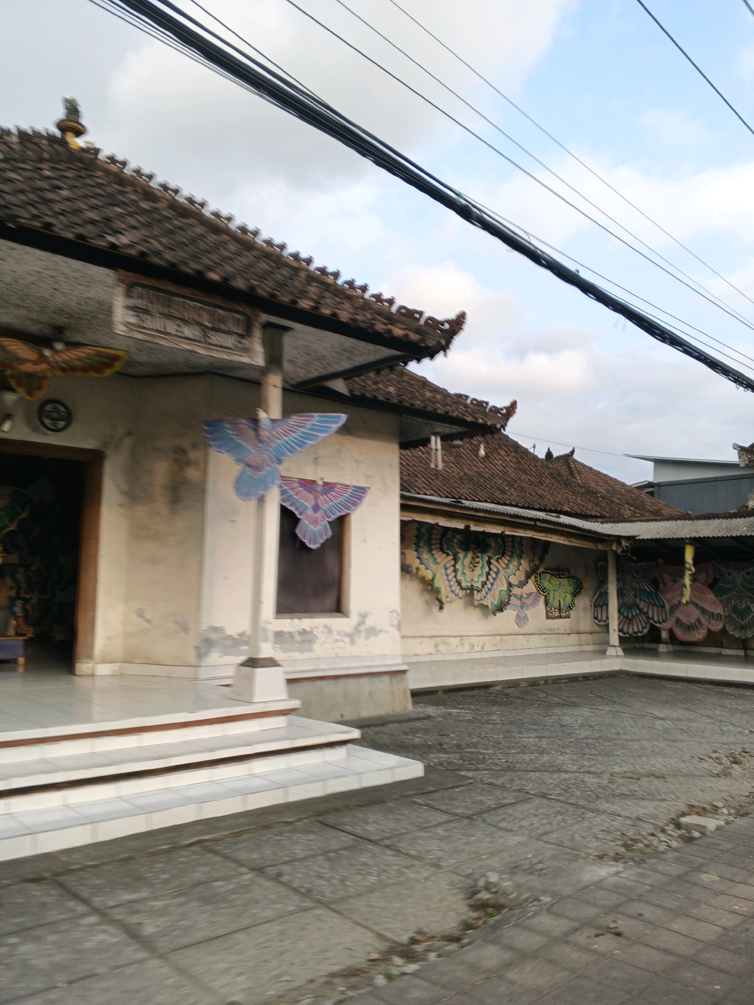 In Sukawati, Indonesia, a scene unfolds in front of a building with a traditional Balinese tiled roof. The building's facade is adorned with colorful, large butterfly-shaped decorations, some hanging from poles and others affixed to the wall. A sign above the entrance appears to read "SOUVENIR SHOP". White tiled steps lead up to the entrance. The ground is paved with gray square tiles. Overhead, a tangle of black electrical wires stretches across the sky, which is a mix of blue and clouds. The overall impression is of a vibrant shop displaying decorative items, likely kites or banners, in an outdoor setting.