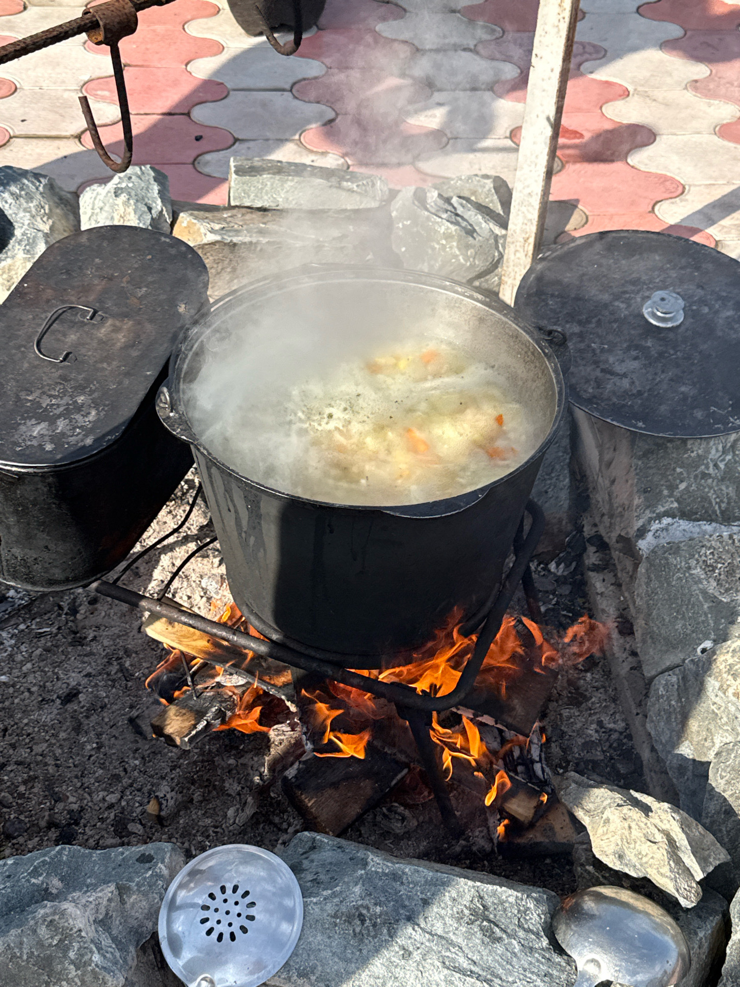 A large black cauldron filled with a bubbling stew is centered in the frame, suspended over a crackling wood fire. The stew contains visible chunks of vegetables, including orange carrots and lighter colored pieces. Steam rises vigorously from the cauldron. To the left of the cauldron, another black pot with a lid is visible, and to the right, a similar pot with a silver knob on its lid. The pots and cauldron are surrounded by rough gray stones. The ground is a mix of dark soil, ash, and small stones, with some larger stones scattered around. In the background, a paved area with interlocking pink and white tiles is partially visible, as is a rusty metal structure with hooks overhead. A metal ladle with a perforated scoop lies on the ground in the foreground to the left. The scene appears to be outdoors, possibly in a rustic setting or an outdoor cooking area. The location is identified as Pakly, Russia. There are no people visible in the image.