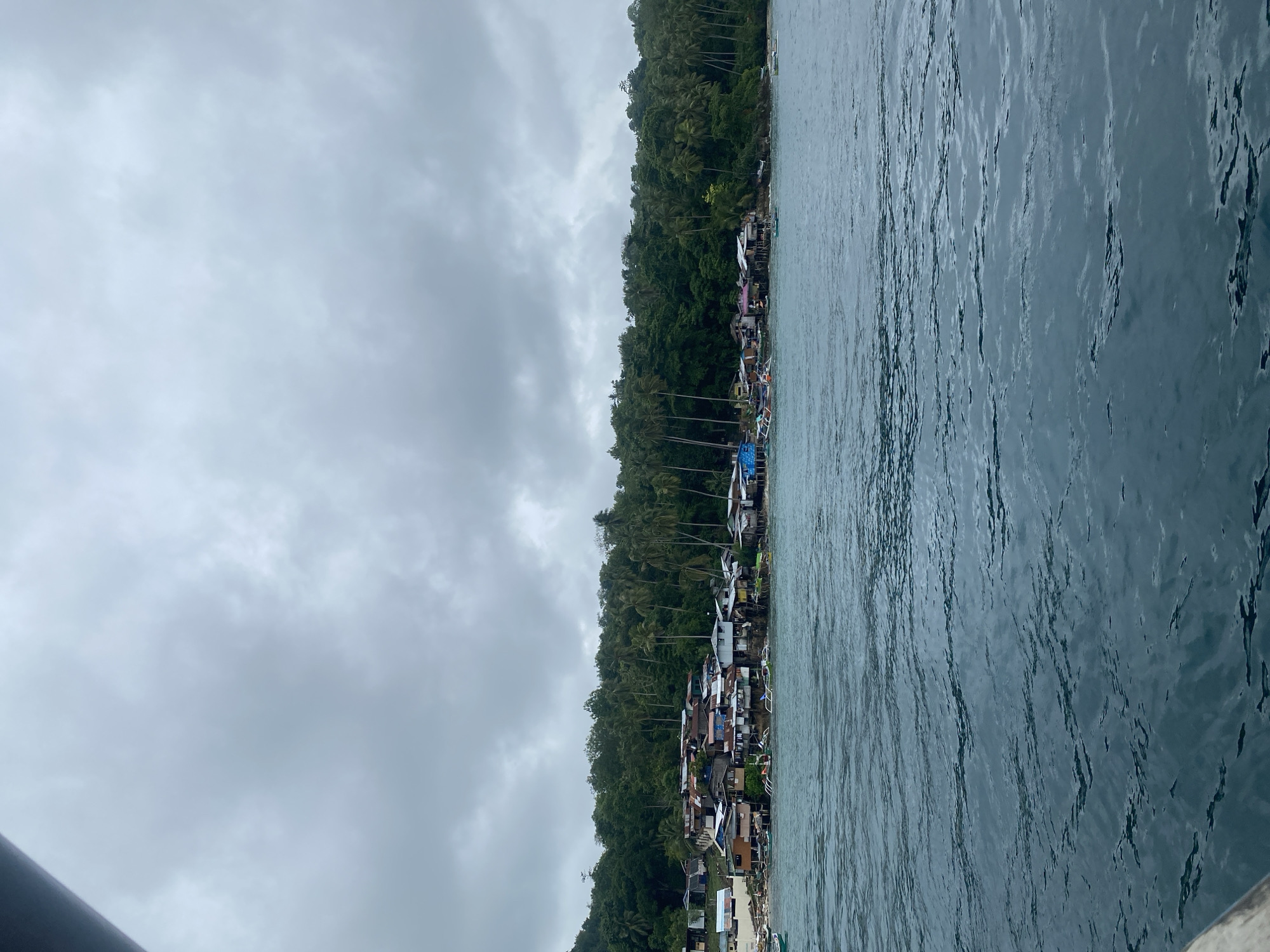 An overhead shot captures a coastal village in Samal, Philippines, under a cloudy sky. The village is nestled between a lush, green, tree-covered hillside and the deep blue, choppy waters of the ocean. Houses, many of them appearing to be built on stilts, are clustered together along the shoreline. The structures are a mix of corrugated metal roofs and wooden walls, some painted in various colors like blue and pink. Numerous small boats, some with outriggers, are anchored near the shore or bobbing in the water. The water itself is a dark, rich blue, reflecting the overcast sky and creating ripples and waves across its surface. The scene depicts a serene, yet rustic, coastal community.
