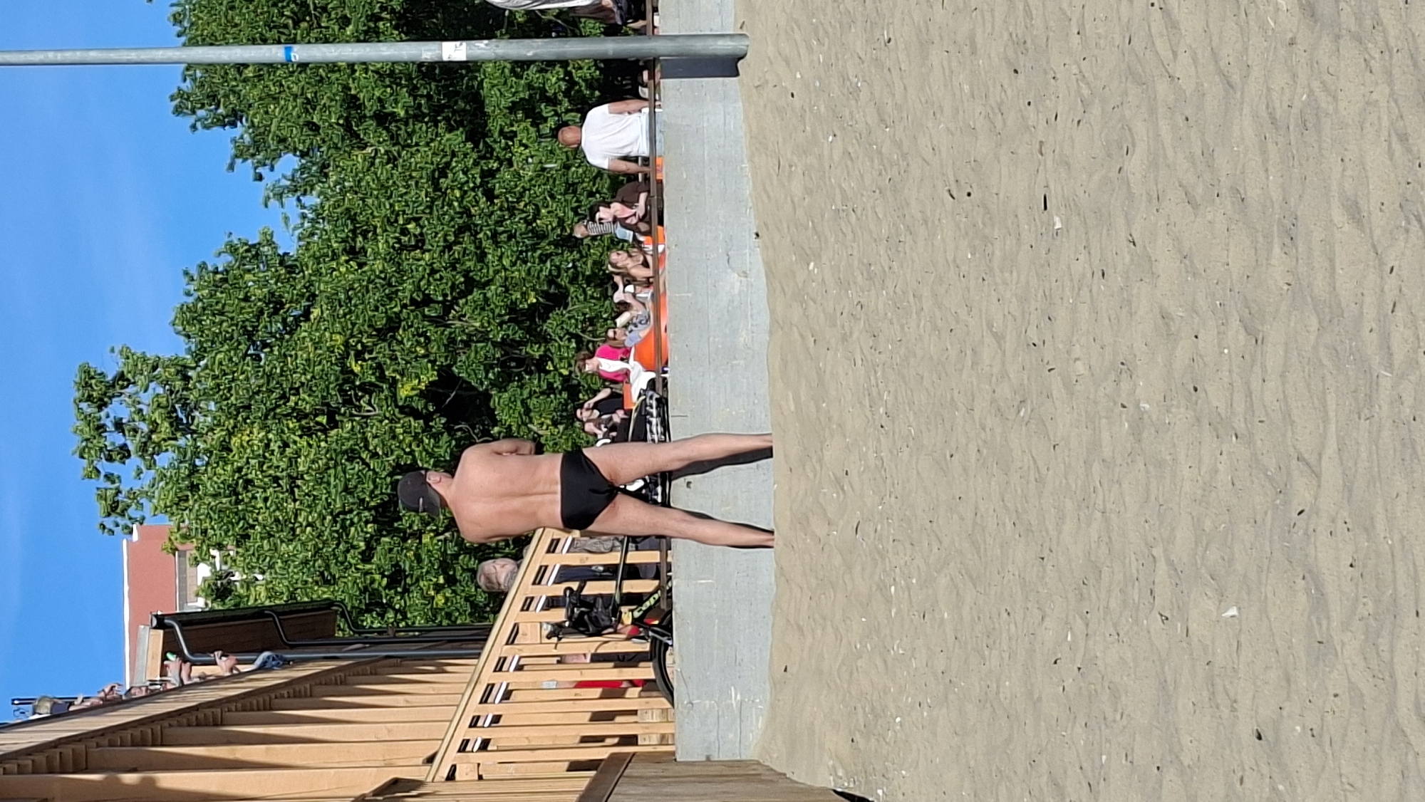 A man wearing black briefs and a black baseball cap stands facing away from the camera on a sandy beach, his back to the viewer. He is positioned on a lower section of the beach, closer to the water. Behind him, a concrete wall separates the sand from a raised area where several people are seated and interacting. Trees and a building are visible in the background. The scene appears to be a public beach on a sunny day. The location is identified as the beach near the Linnahall in Tallinn, Estonia, which is a former ice arena and concert hall situated on the Tallinn Bay waterfront, west of the city center.
