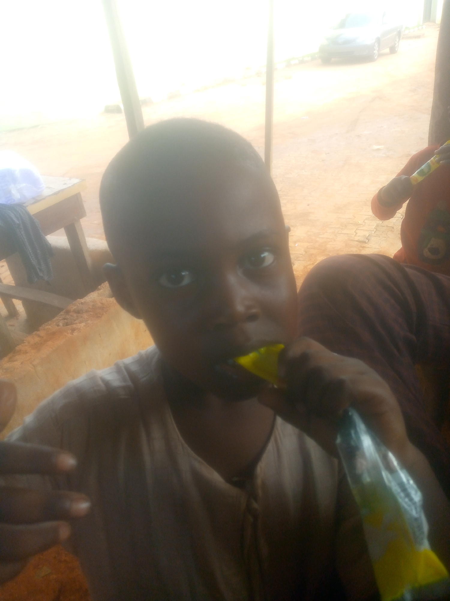 A child is seen in the foreground, holding a yellow and transparent packaging in their mouth. There is a person in the background with a red shirt, wearing brown pants and a red teddy bear design. There is a table on the left side of the image. A light-colored vehicle is parked in the background. The location appears to be outdoors in an area with light-colored dirt or sand. The city and country are Unknown.