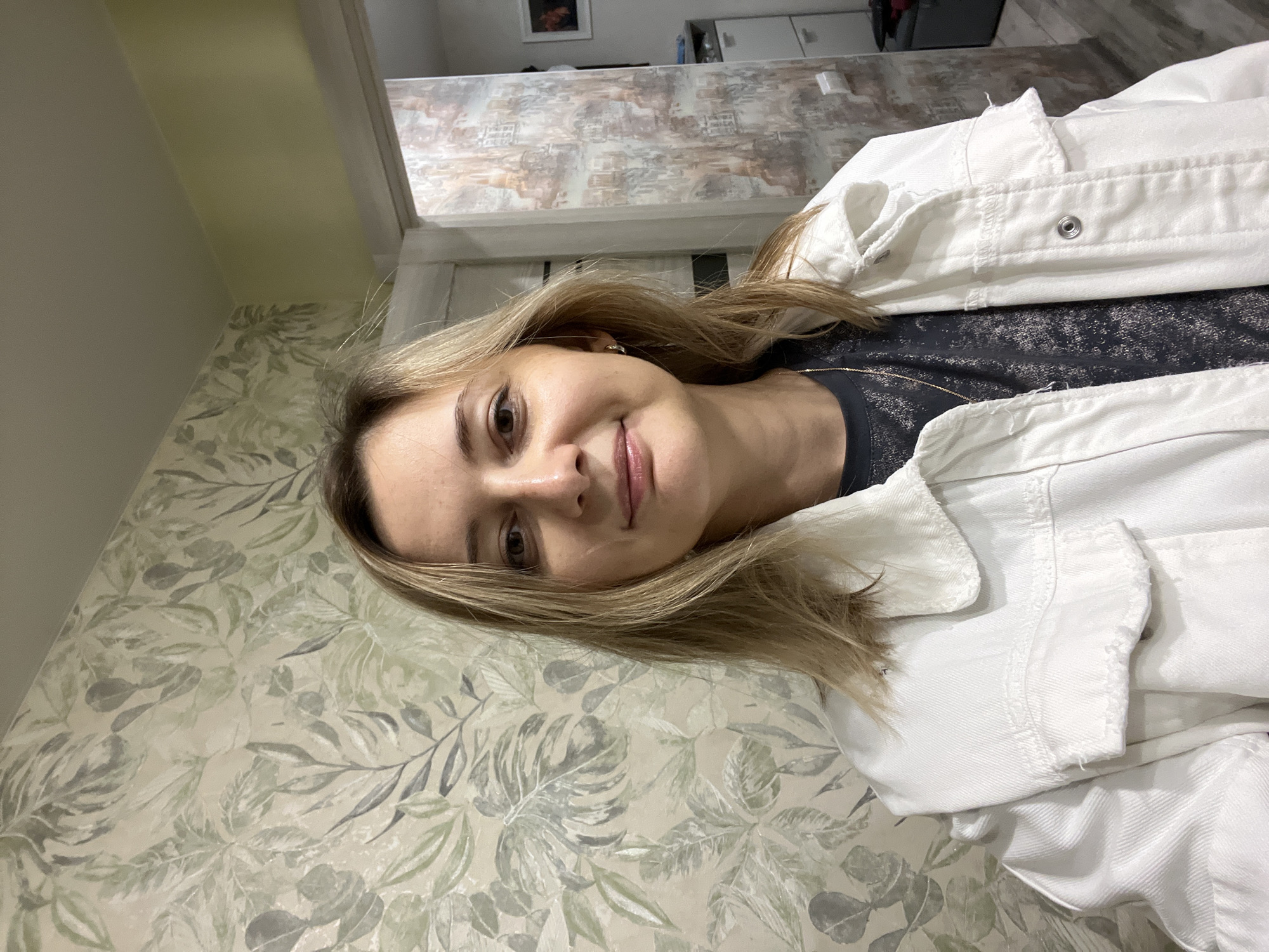 A woman with blonde hair, wearing a white denim jacket and a dark shirt, is looking at the camera with a slight smile. She is in a room with patterned wallpaper. The image is taken from a high angle, and the woman is positioned in the center of the frame. The lighting suggests it is daytime. The wallpaper has a botanical print with leaves in shades of green and grey. Behind the woman, a wall with a different pattern and a doorway are visible. There is no visible text in the image. The setting appears to be a domestic interior in Volgograd, Russia.