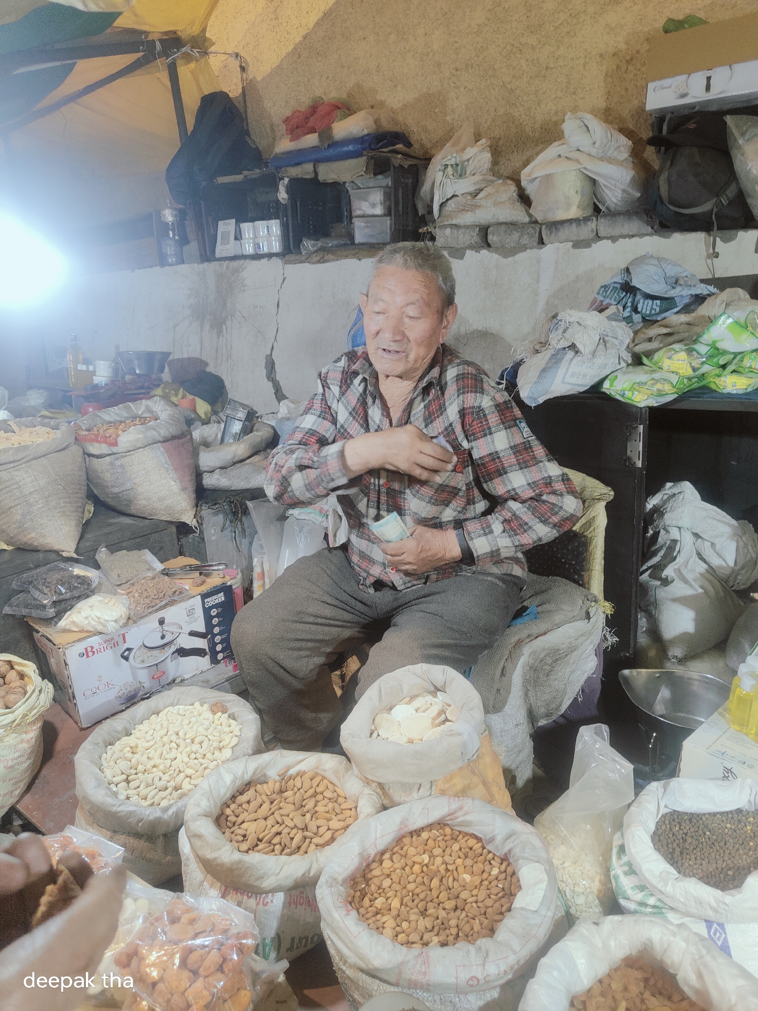 An elderly East Asian man sits in a dimly lit stall in Leh, India, surrounded by sacks of nuts and dried fruits. He is wearing a red and black checkered shirt and dark pants, and he holds a few banknotes in his hand, appearing to be making a transaction. The stall appears to be an outdoor market, possibly a roadside vendor, with a makeshift canopy overhead and concrete walls behind. Sacks of various nuts and dried fruits, including almonds, cashews, and what appear to be raisins, are piled around him. A box with an image of a pressure cooker is also visible. The overall scene depicts a common sight in the bustling markets of Leh, where vendors sell local produce and goods.
