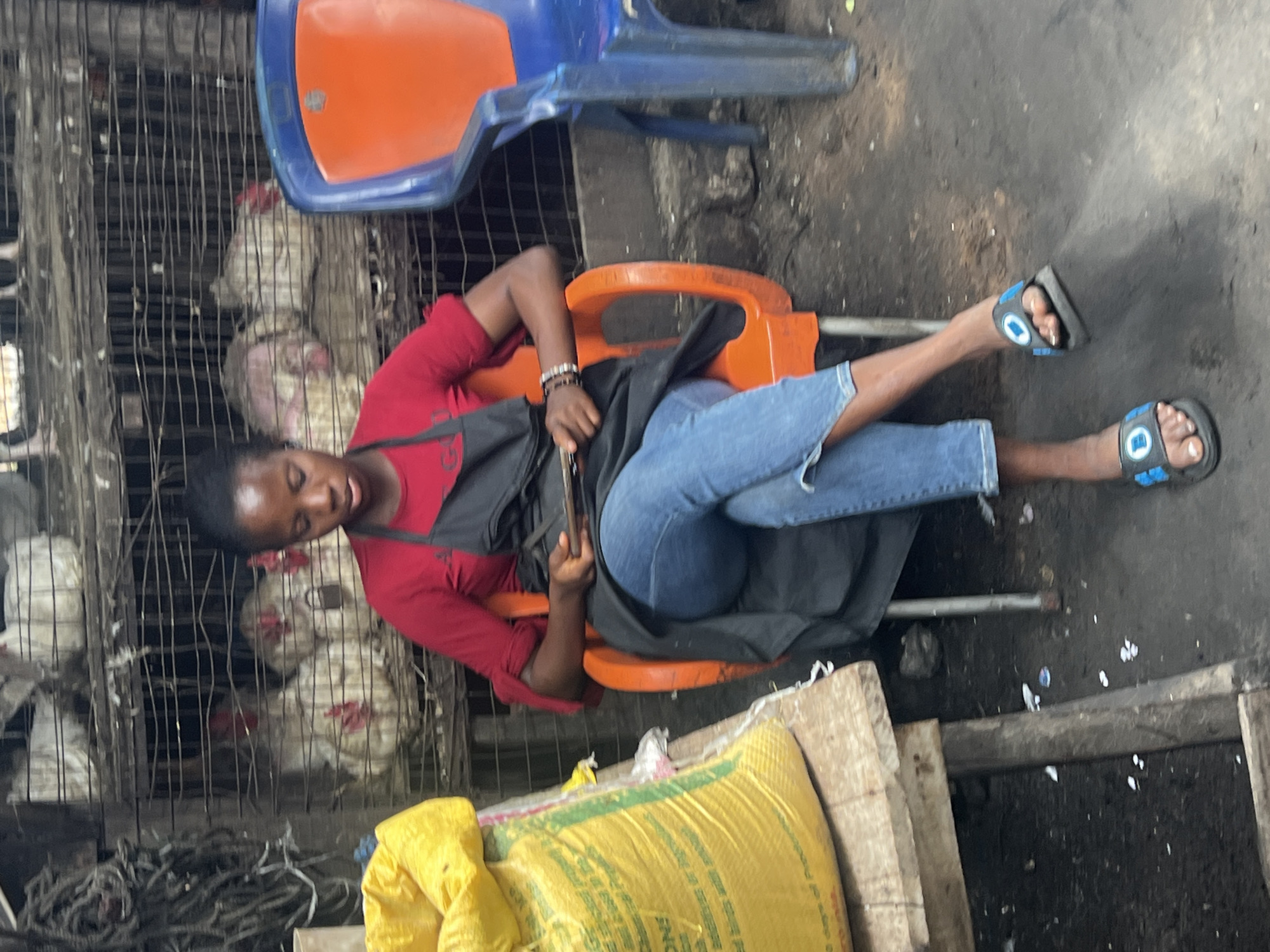 A young woman is seated in an orange plastic chair in front of cages filled with chickens, likely at a market in Nkpor, Nigeria. She is wearing a red t-shirt, blue jeans, and a black apron. She is looking down at a mobile phone in her hands. To her left is a large yellow bag, possibly containing feed, and to her right is a blue plastic chair. The ground appears to be dirt and debris. The chickens are in wire cages stacked on shelves. The overall scene depicts a market environment where live poultry is sold.