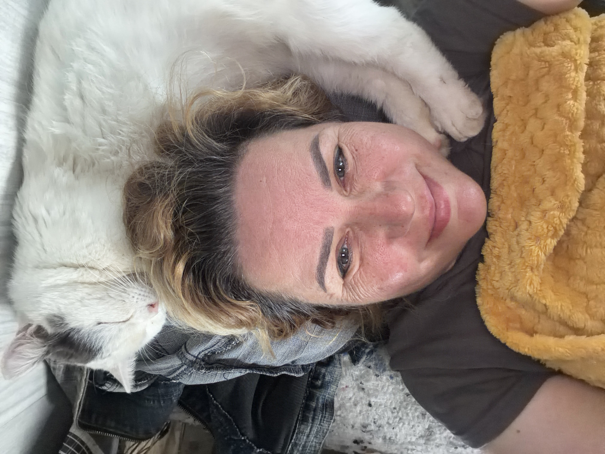 A woman is lying on her back, smiling at the camera. A white and grey cat is lying on her chest, mostly asleep. To her right is a yellow fuzzy blanket. She appears to be indoors on a bed. It is not possible to determine if this is located in Leningradskaya, Russia from the given image.