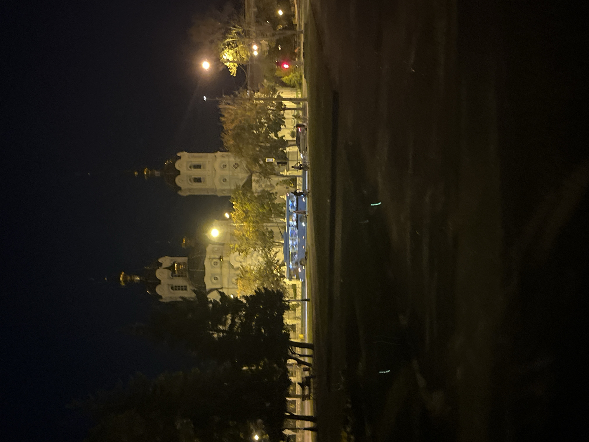 The image depicts a nighttime scene in Kharkiv, Ukraine, featuring the Dormition Cathedral. The cathedral's distinctive white facade and golden domes are illuminated against the dark sky, with its spires reaching upwards. In the foreground, silhouetted trees cast shadows, and a street scene unfolds. A blue bus is visible on the road, with its headlights on. Several individuals are also present, some on bicycles and others walking. The environment appears to be a public space with streetlights casting a warm glow on the surroundings. The overall impression is a quiet evening in the city, with the grand cathedral serving as a prominent landmark.