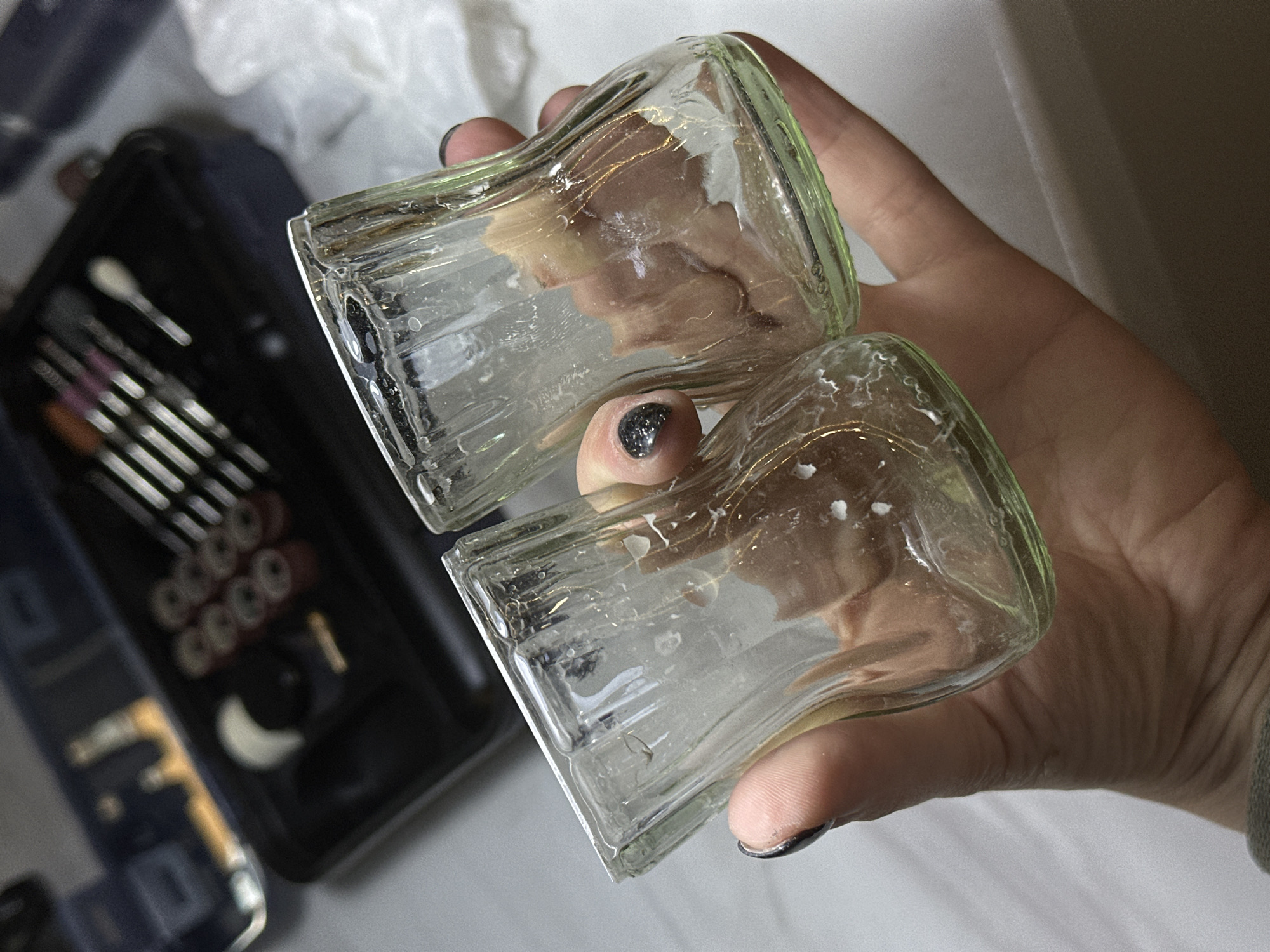 A close-up, slightly angled indoor shot shows a person's hand holding two small, clear glass bottles with a subtle light green tint. The bottles are rectangular in shape with fluted sides and appear to be stuck together or held very tightly. White, possibly dried, residue is visible on the interior and exterior surfaces of the glass. The person's thumb and index finger are visible, with dark, glittery nail polish on the fingernails.
In the blurred background to the left, a black case is open, revealing a collection of tools, likely makeup brushes and applicators, arranged neatly. Several small, circular, metallic objects, possibly rings or beads, are also indistinctly visible within the case. The lighting is bright, suggesting an indoor setting. There is no visible text in the image. The scene is set in Kuda, Russia.