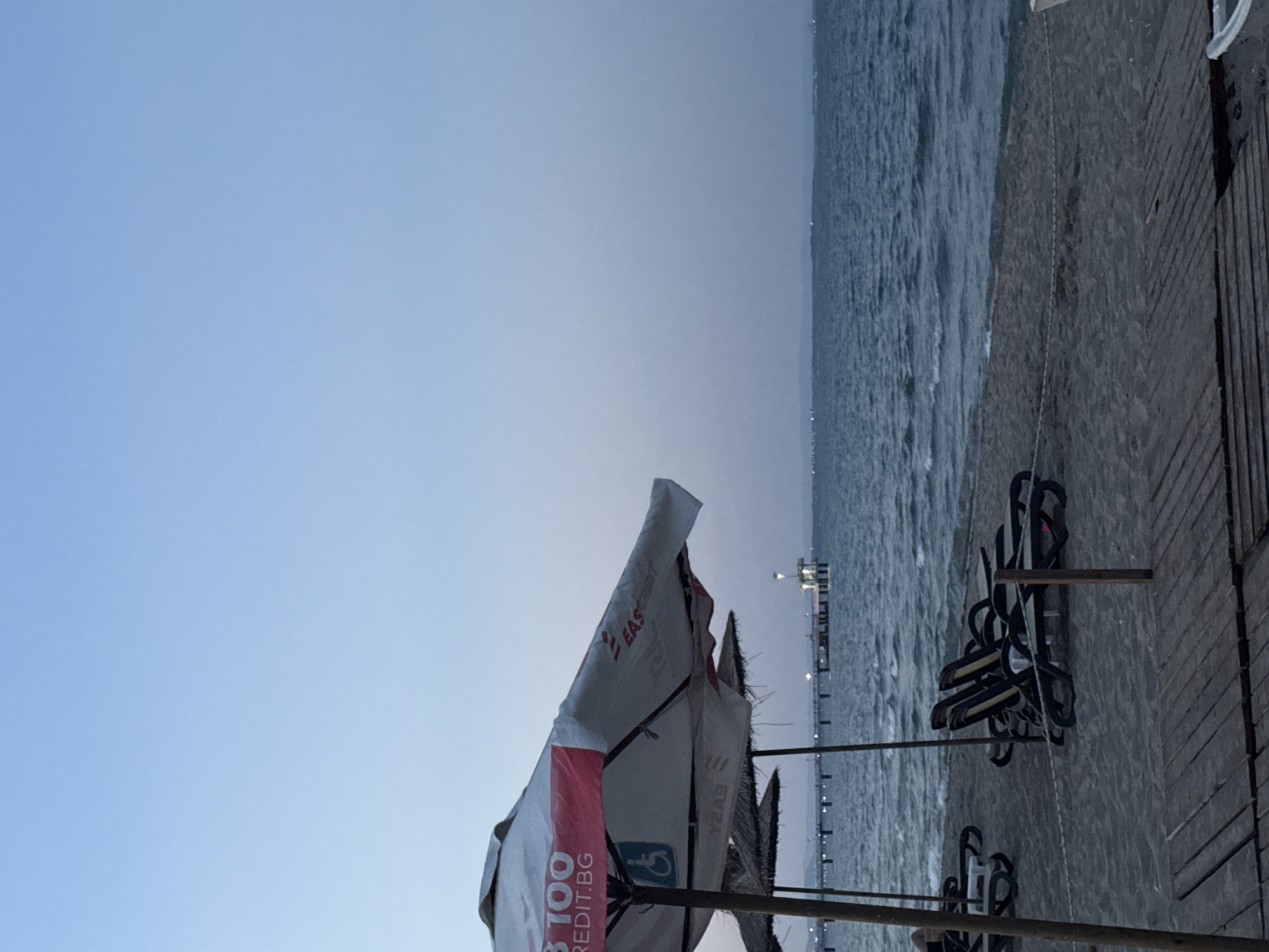 The image depicts a serene beach scene at dusk in Burgas, Bulgaria. In the foreground, a wooden walkway leads towards a sandy beach where several empty lounge chairs and umbrellas are set up. Beyond the beach, the calm, dark blue waters of the sea stretch towards the horizon, where a long pier extends into the water, illuminated by several lights. The sky is a pale, hazy blue, typical of twilight. The overall atmosphere is peaceful and quiet, suggesting the end of a day at the beach.