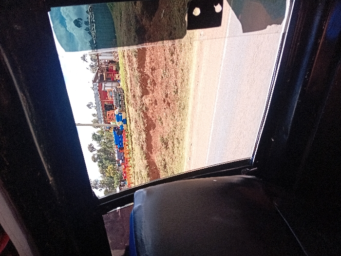 The image depicts an outdoor scene viewed from inside a vehicle, through a side window. In the foreground, the dark interior frame of the vehicle and a portion of a light-colored seat are visible. A paved road, light grey with a faint white line, extends across the lower mid-ground. Beyond the road, a raised bank of reddish-brown soil forms the immediate roadside.
In the background, an open-air commercial or storage area is situated on an elevated plot. This area contains numerous stacked items, including bright blue and red plastic drums or barrels, and multiple rows of red objects resembling plastic chairs or crates. A red-roofed building, constructed from corrugated metal or similar materials, stands centrally within this area, flanked by other structures and fences. Several indistinct human figures are discernible near these structures, indicating activity. The distant background features a dense aggregation of tall, slender trees. The sky is bright and mostly clear, with scattered white clouds, one of which is prominently reflected on the vehicle's windowpane on the right side. The scene is located in Kisima ward, Kenya.