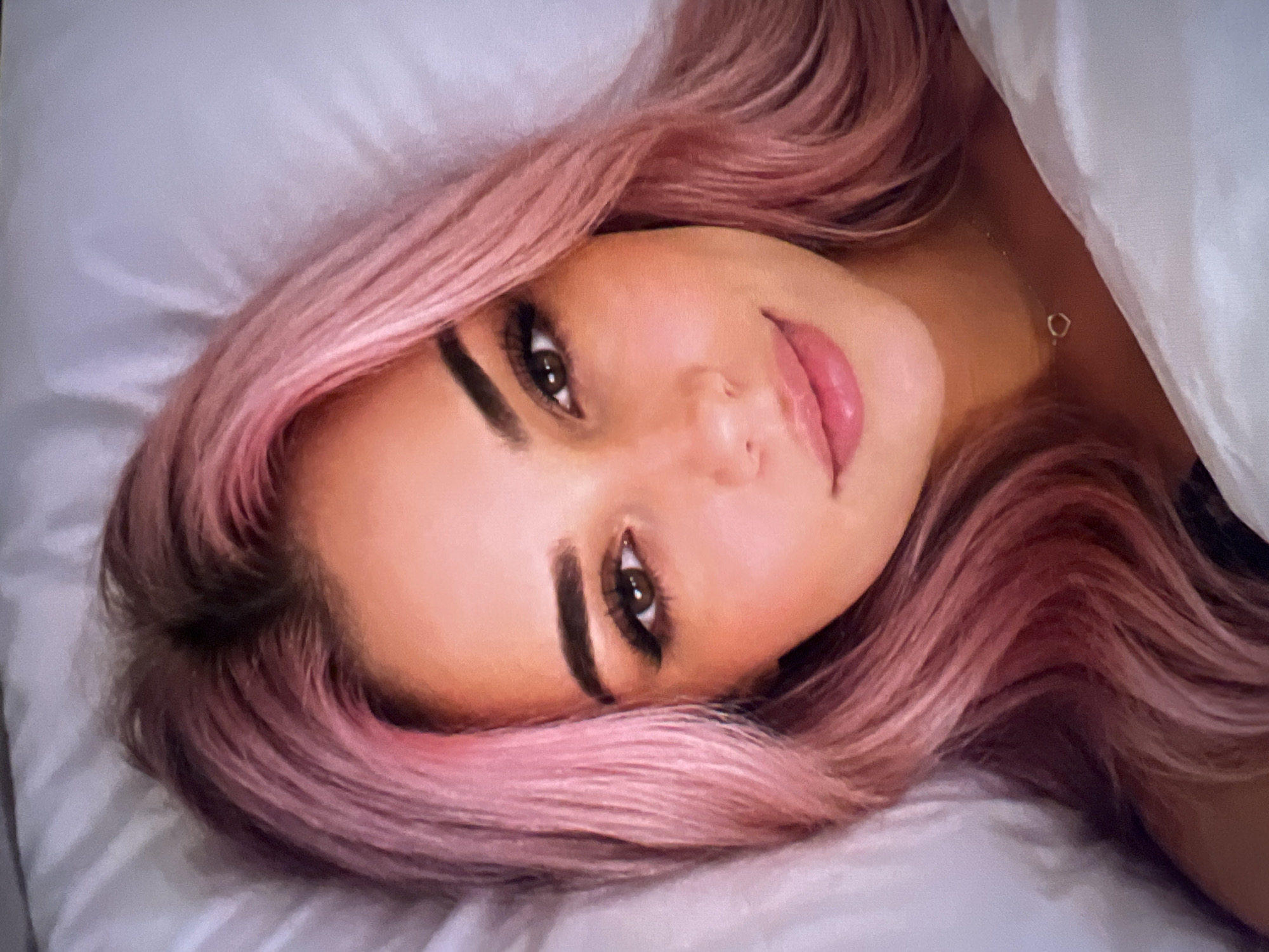 A close-up, head-and-shoulders shot shows a young woman with pink hair lying on a white pillow. Her hair is parted in the middle and falls over her shoulders. She is wearing a dark top and a thin necklace with a small heart pendant. Her eyes are brown and she has on dramatic winged eyeliner and false eyelashes. Her lips are full and painted with a glossy pink lipstick. The background is blurred and appears to be a white sheet or duvet. The lighting is soft and warm.