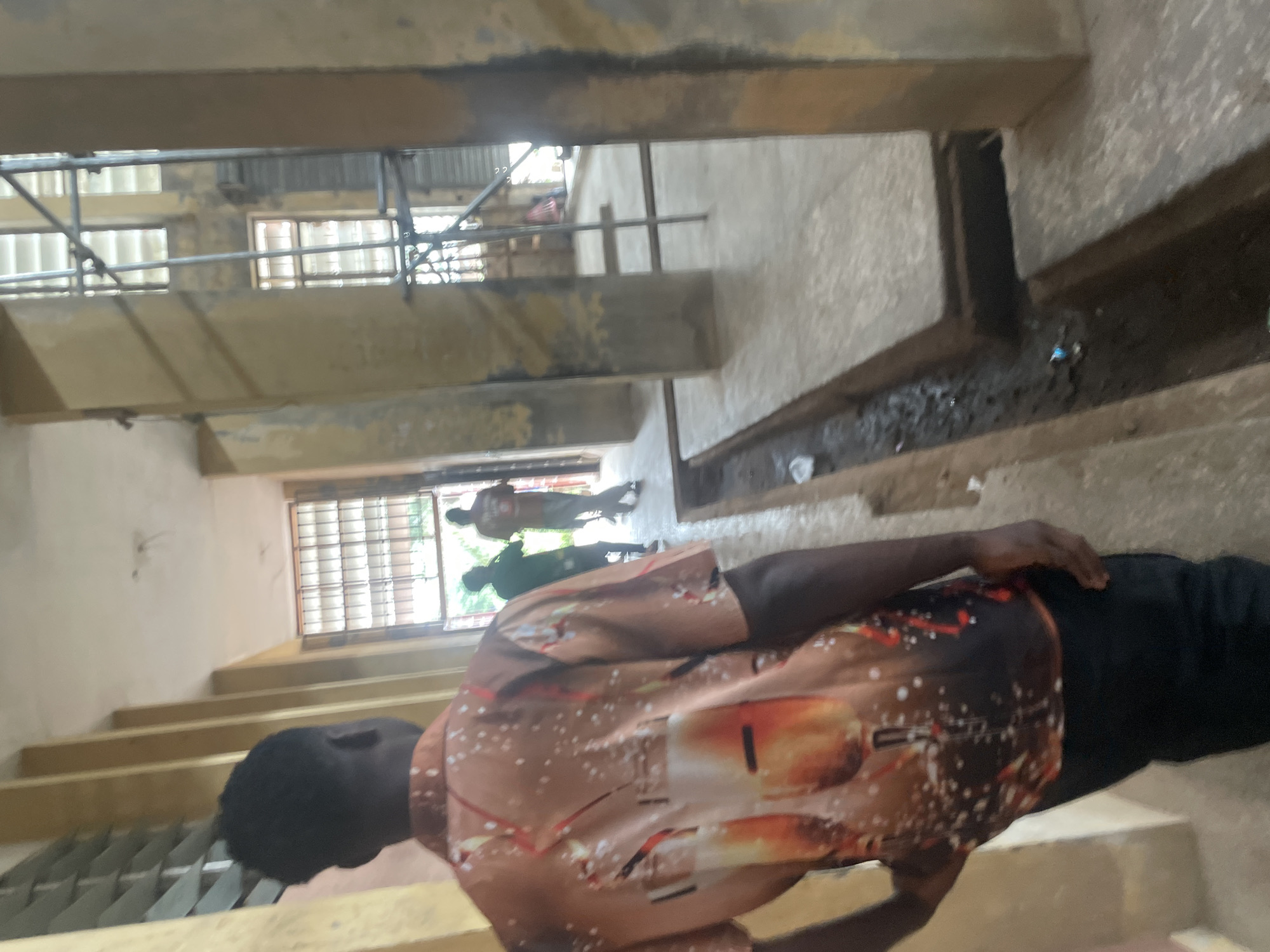 The image depicts an outdoor hallway in what appears to be a school or institutional building in Ihiagwa, Nigeria. In the foreground, a Black male wearing a patterned short-sleeved shirt with images of glasses and a dark-colored pair of pants is seen from behind, walking away from the camera. His hair is short and dark. Further down the hallway, two other individuals are visible. One person is wearing a green shirt and the other a light-colored shirt. They are walking in the same direction as the person in the foreground. The hallway is flanked by concrete pillars and walls with a yellowish-brown hue. There are barred windows on the left side of the hallway and a metal scaffolding structure on the right. The floor of the hallway appears to be concrete, with a visible drainage channel running along the right side. The lighting suggests it is daytime, with light filtering in from openings at the end of the hallway. The overall environment appears somewhat worn and utilitarian.