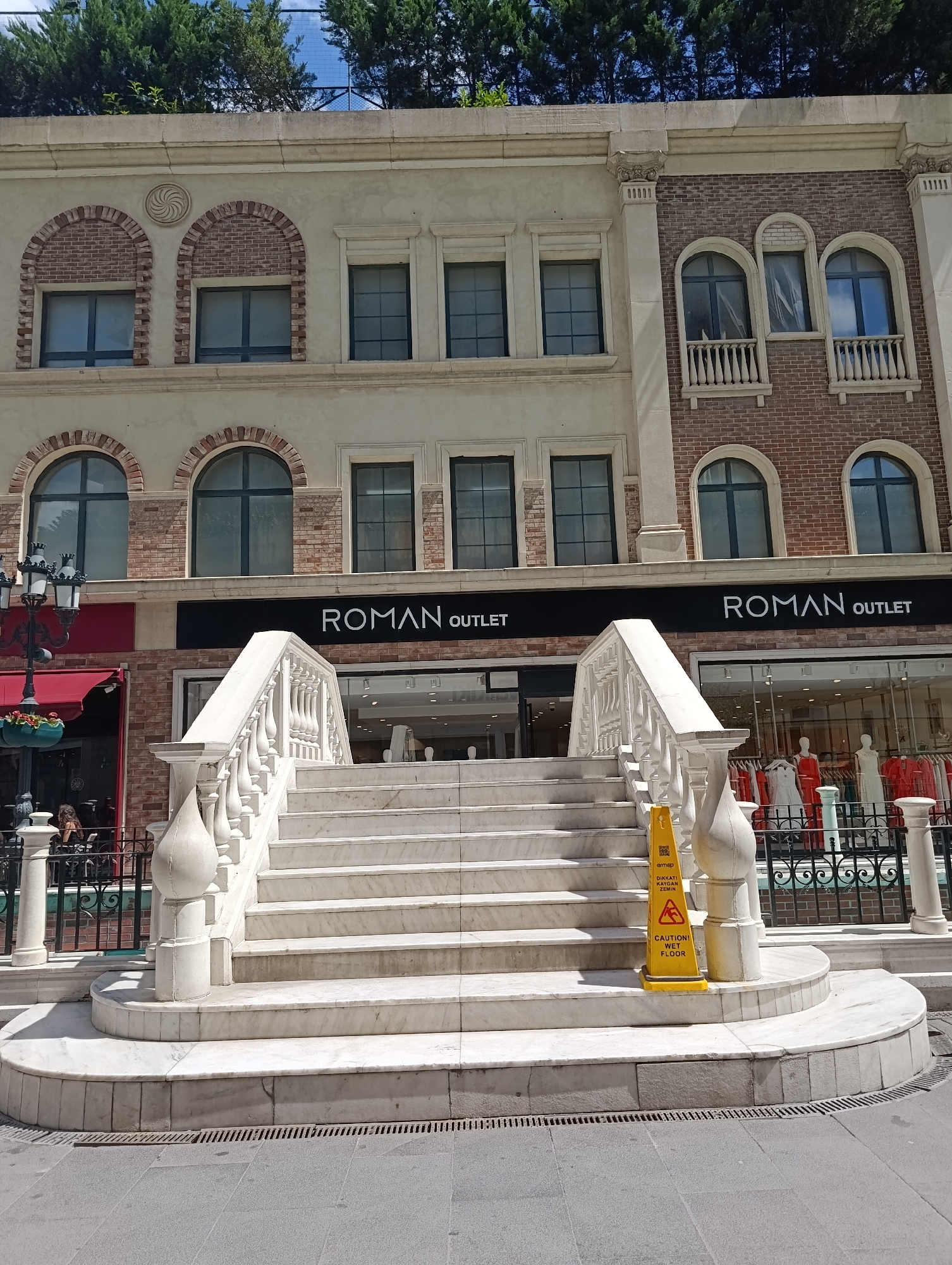 The image shows the entrance to a "Roman Outlet" store located in Gaziosmanpaşa, Turkey. A grand white marble staircase leads up to the store's entrance. A yellow caution cone, with "CAUTION WET FLOOR" written on it, is placed on the staircase. The store's facade features a mix of architectural styles, with arched windows and brickwork. Green trees are visible on the rooftop. Inside the store, mannequins are displayed with clothing. In the foreground, a person is seated at an outdoor cafe to the left of the stairs. The street is paved with grey tiles and features a drainage grate.